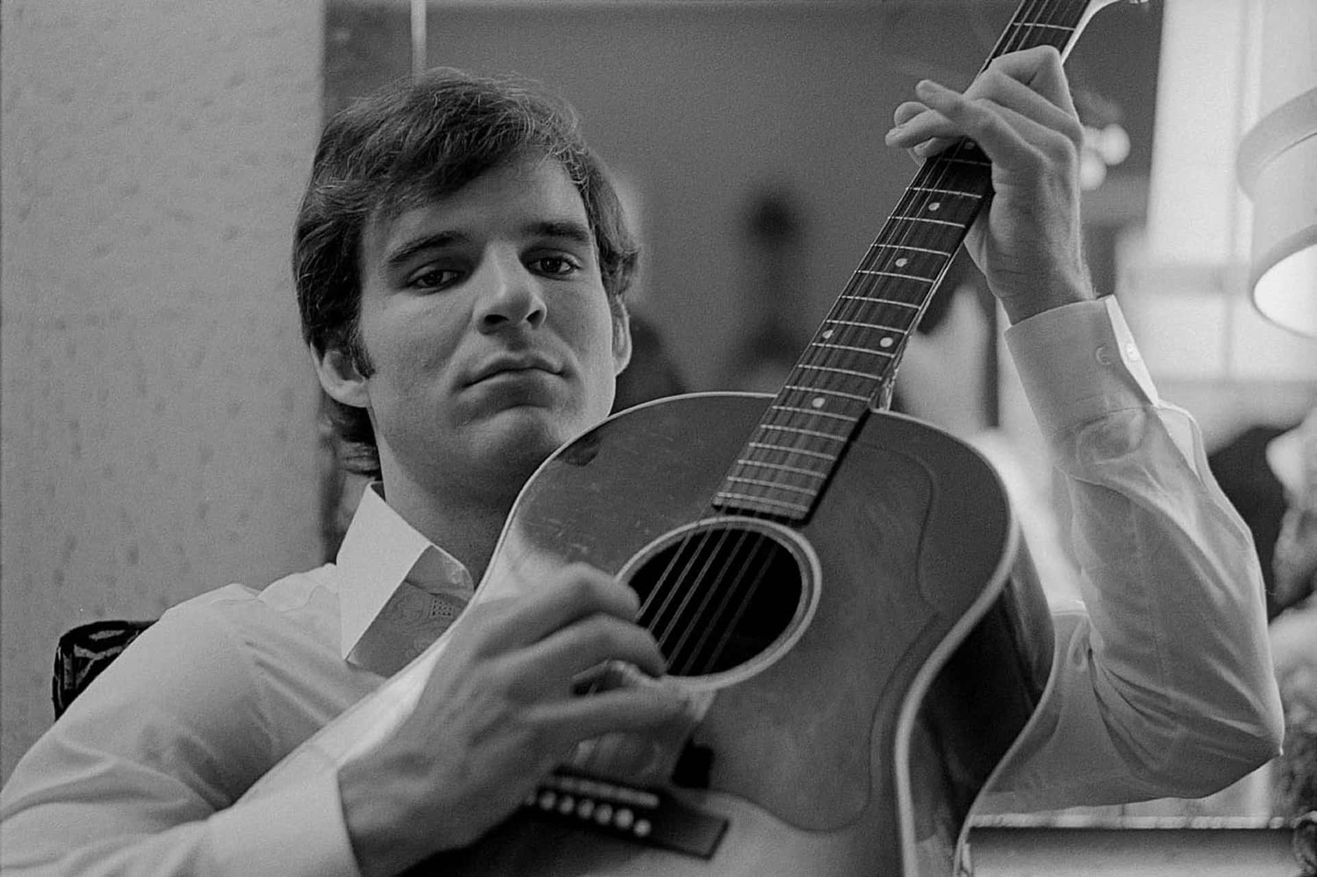 steve martin 桌布 壁紙 — 一位穿著有領襯衫的年輕男子在柔和光線的室內環境中，專注地抱著一把原聲吉他。