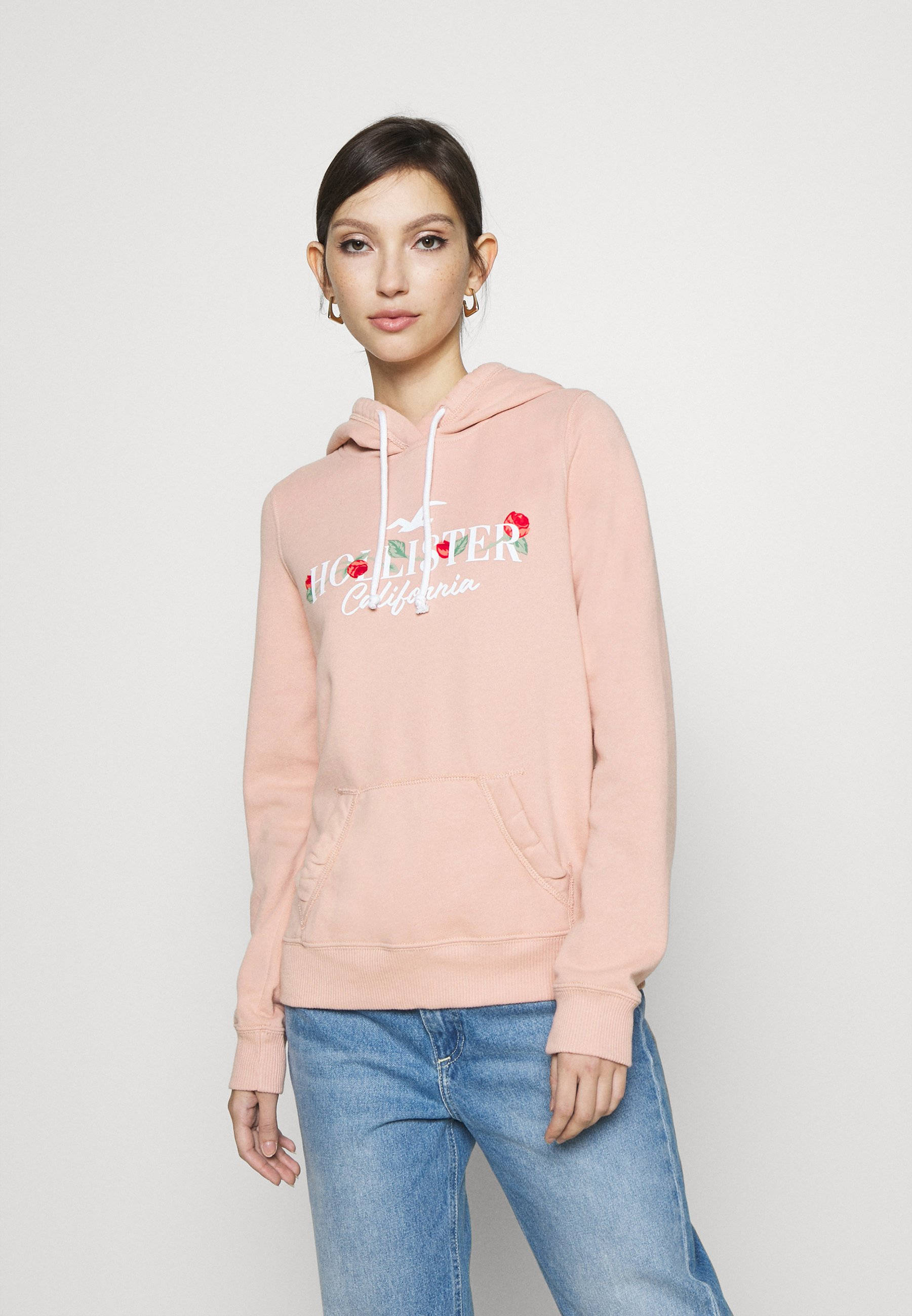 Hollister 桌布 壁紙 — 一位穿著柔軟粉紅色連帽衫（帶有櫻桃刺繡）和藍色牛仔褲的女性在純白色背景前擺姿勢。