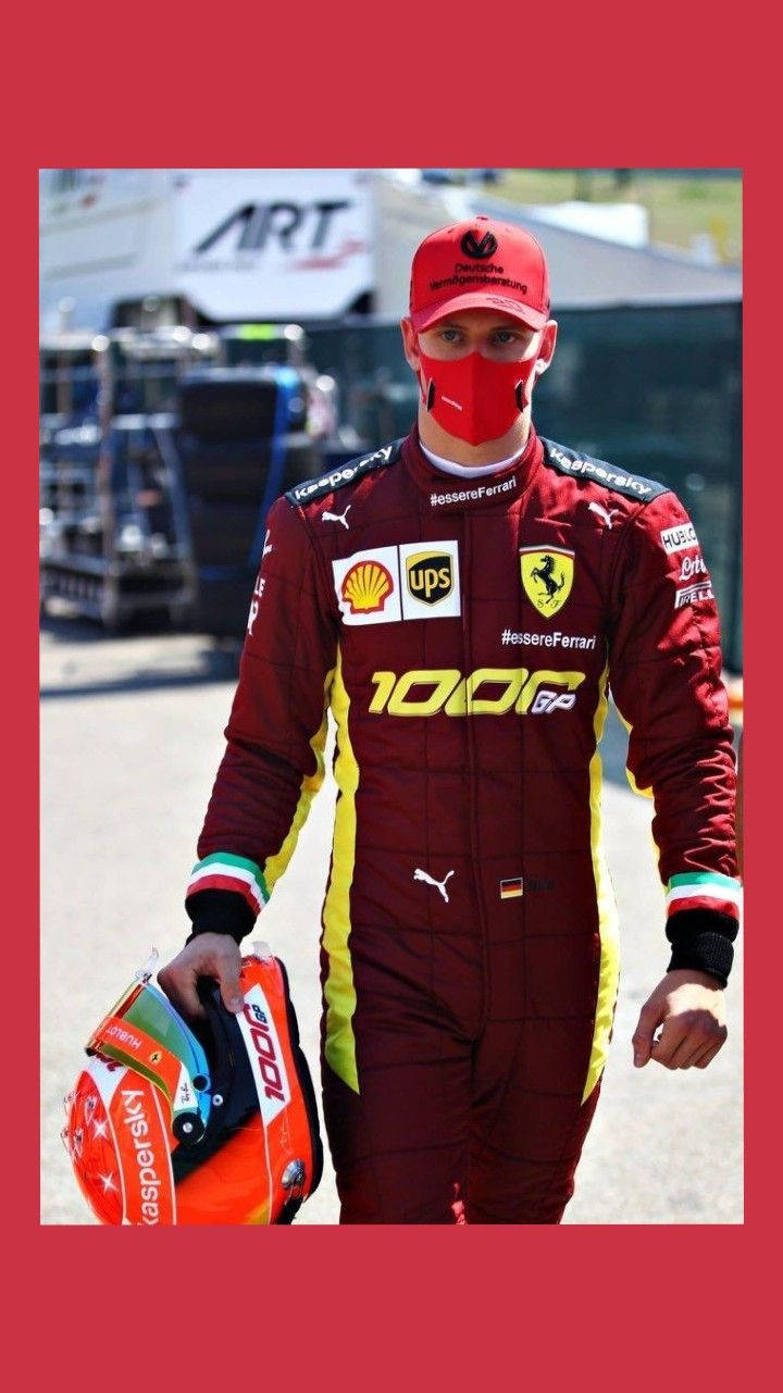 mick schumacher 桌布 壁紙 — 一位穿著栗色法拉利賽車服和紅色口罩的一級方程式車手，在陽光明媚的賽道上拿著頭盔行走。