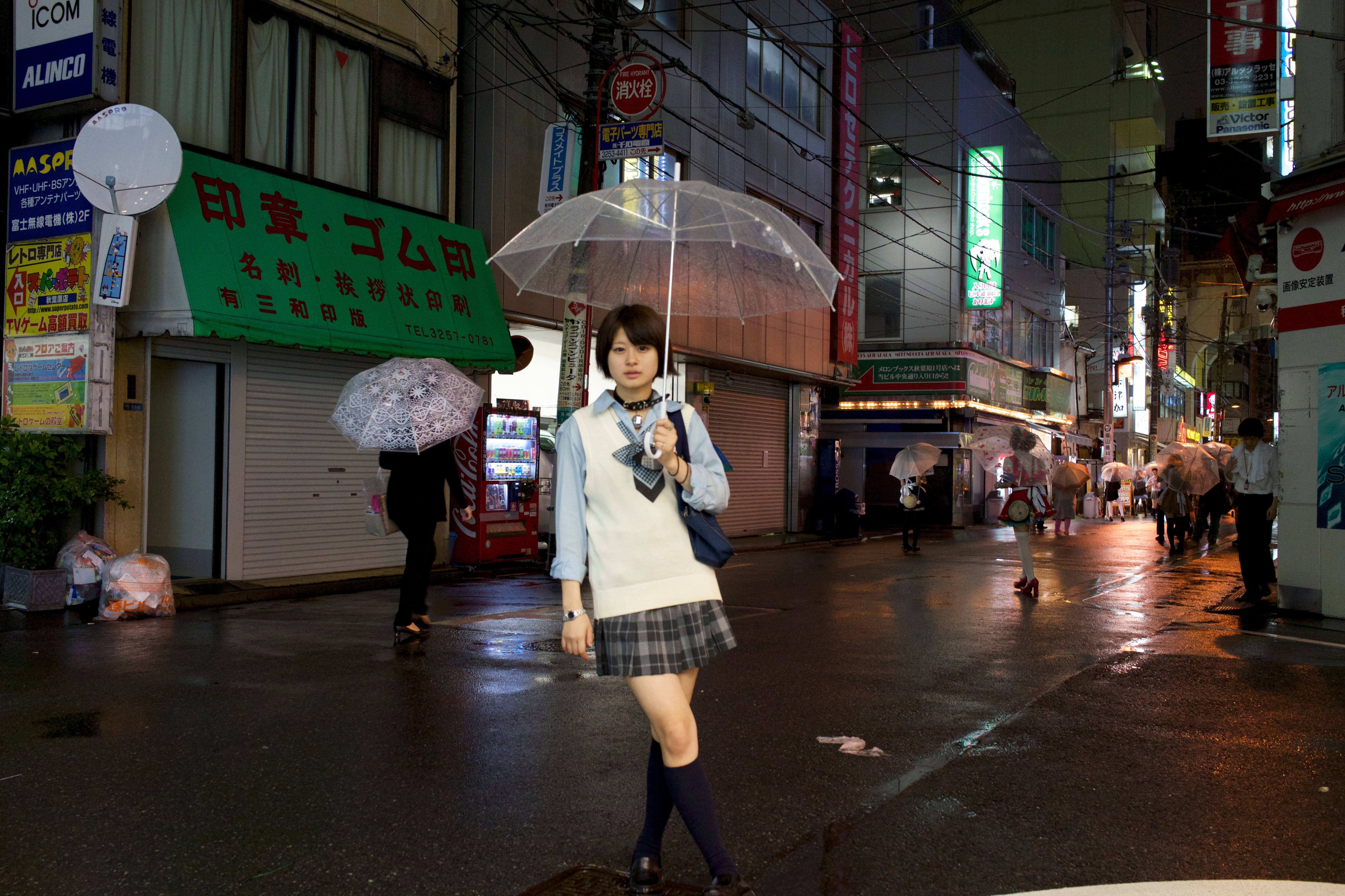 Raining 桌布 壁紙 — 一位穿著校服的年輕女性自信地走在霓虹燈閃爍的雨夜東京街道上，手持透明雨傘。