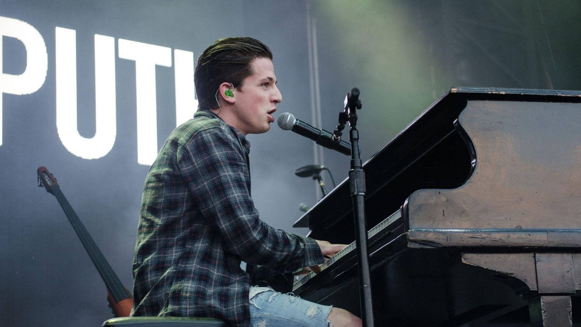 Charlie Puth 桌布 壁紙 — 一位穿著格子襯衫的音樂家在明亮的舞台燈光下，身後有一個大螢幕，正在彈奏平台鋼琴。