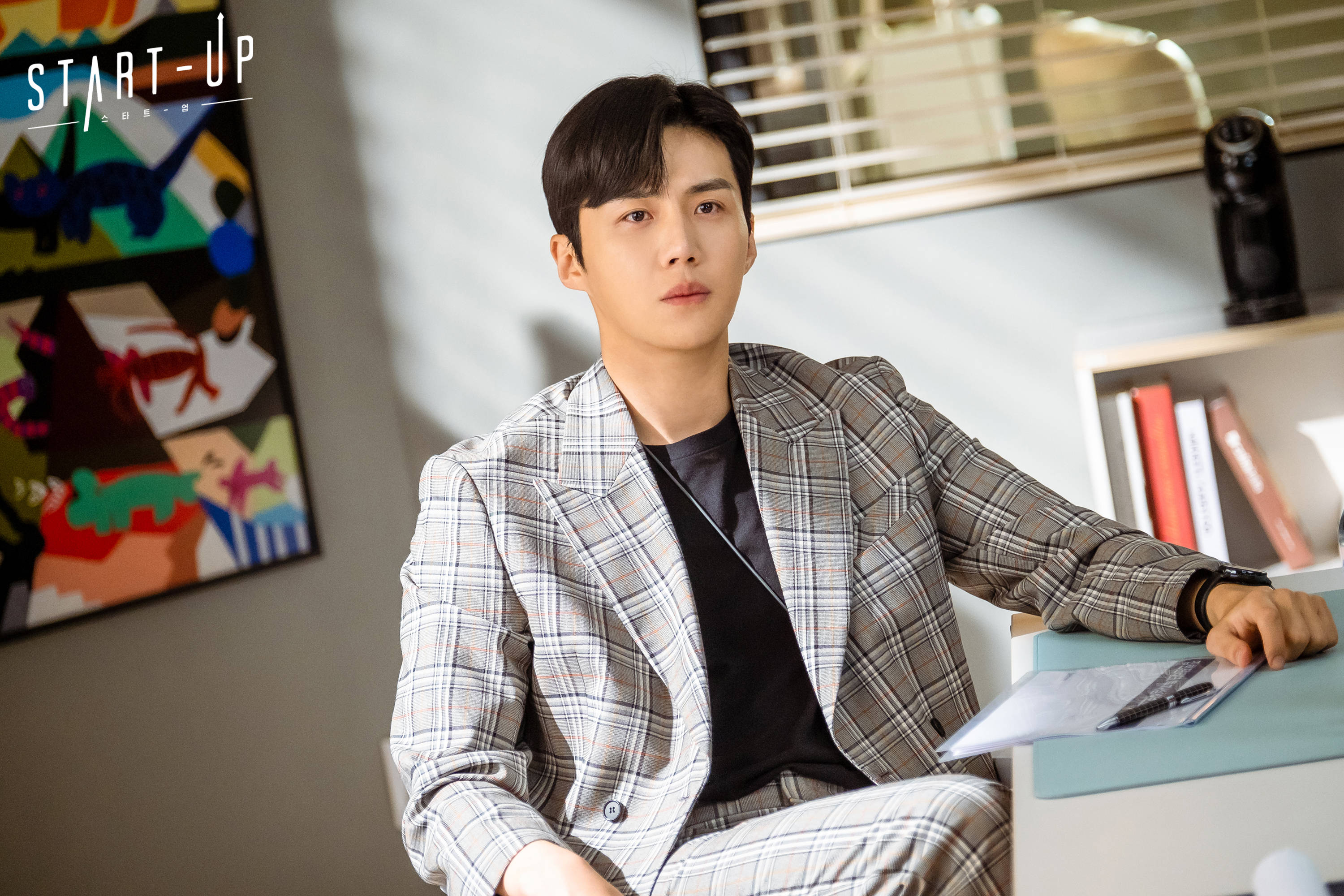 Kim Seon Ho 桌布 壁紙 — 一位穿著格子西裝的年輕男子坐在現代辦公室的辦公桌前，牆上掛著抽象藝術品，若有所思。