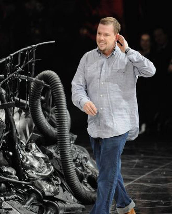Celebrity 桌布 壁紙 — 一位穿著格紋襯衫、面帶微笑的男子，在光線昏暗的室內空間，走過一個帶有螺旋管的大型金屬雕塑。