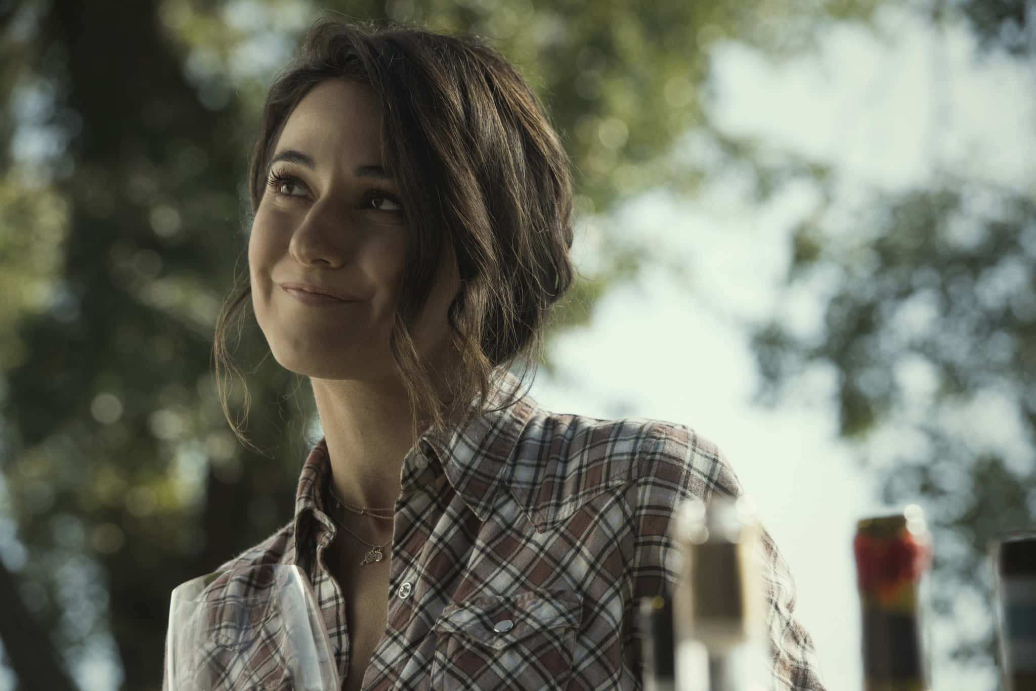 Emmanuelle Chriqui 桌布 壁紙 — 一位穿著格紋襯衫的年輕女性，在戶外樹木間灑落的斑駁陽光下溫柔微笑。