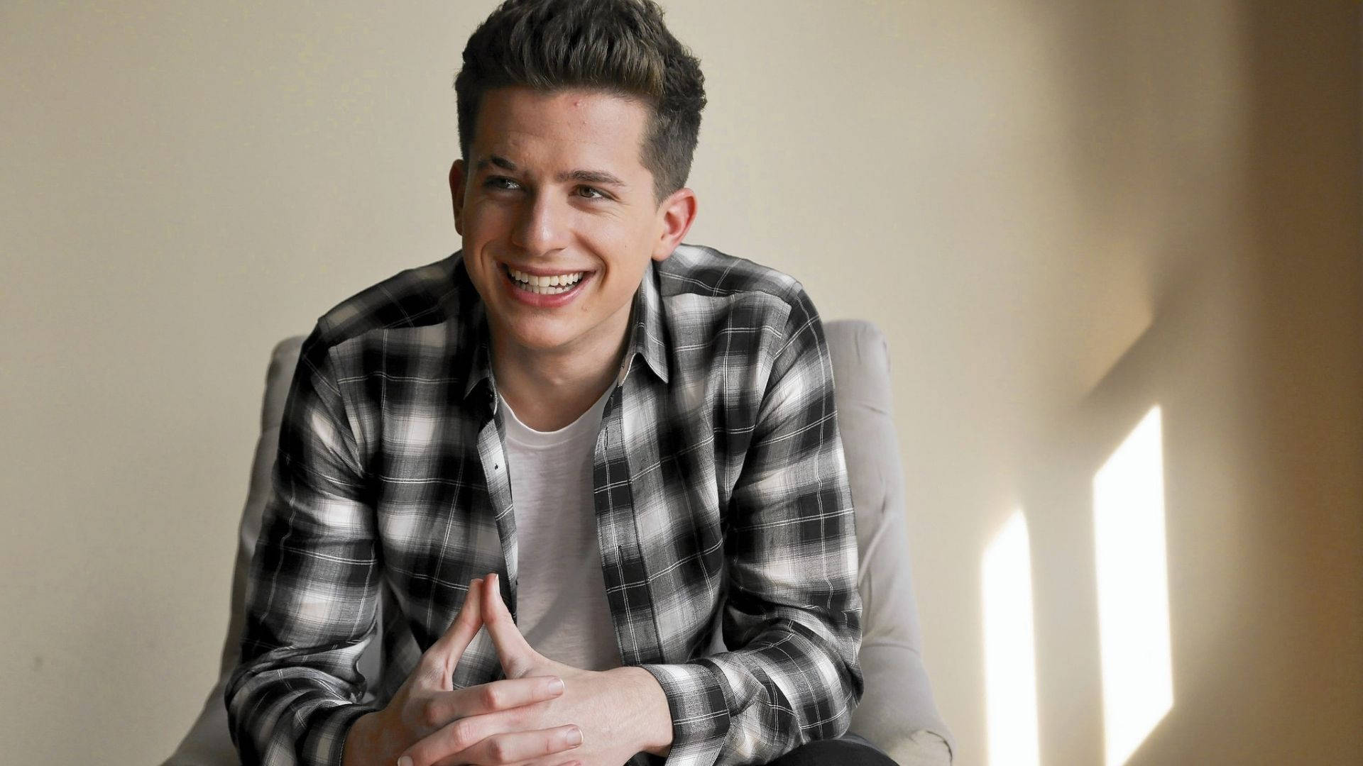 Charlie Puth 桌布 壁紙 — 一位穿著格紋襯衫的年輕男子，坐在柔和光線的房間裡，陽光從窗戶灑進來，露出溫暖的笑容。