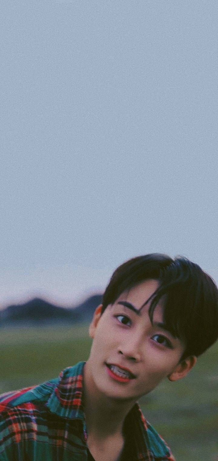 yoon jeonghan 桌布 壁紙 — 一位穿著格紋襯衫的年輕男子在暮色山景背景下柔和地微笑，色調為柔和的藍色和綠色。
