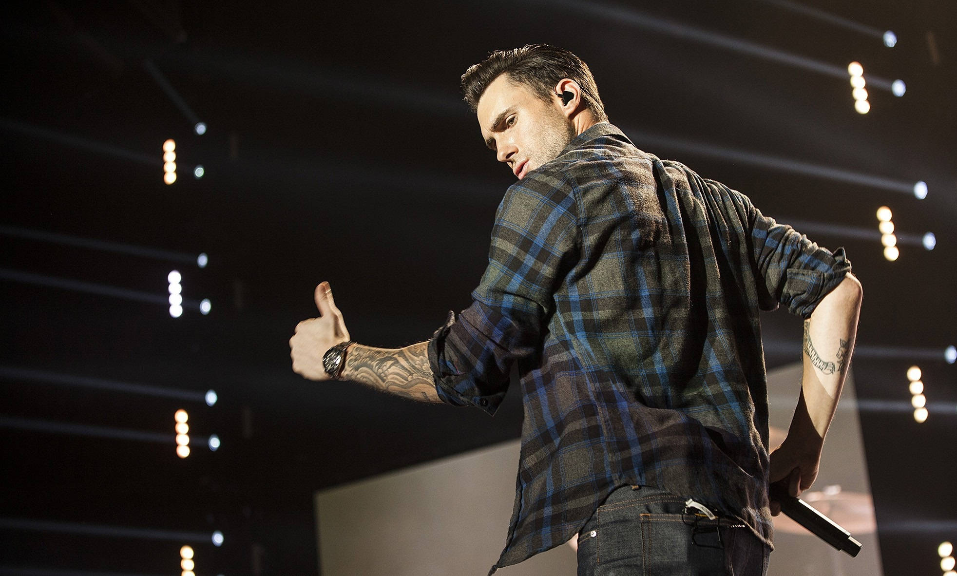 Adam Levine 桌布 壁紙 — 一位穿著格紋襯衫的男表演者在燈光昏暗的舞台上，在聚光燈下豎起大拇指。