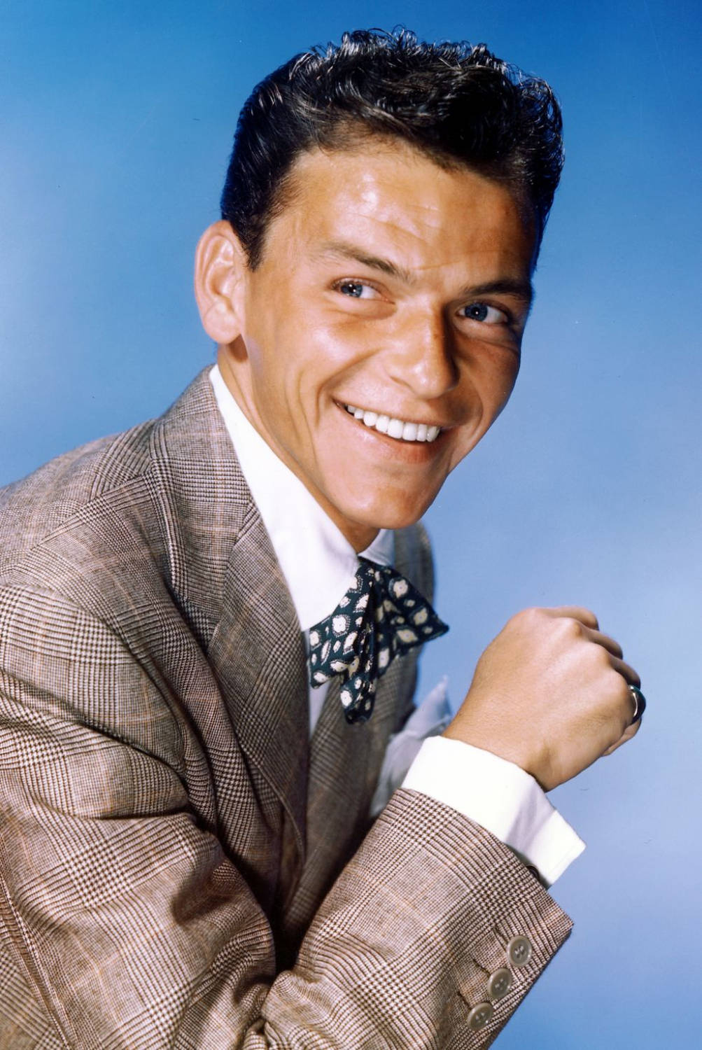 Sinatra 桌布 壁紙 — 一位穿著格紋西裝和圖案領結的開朗男子，在純藍色攝影棚背景下燦爛微笑。