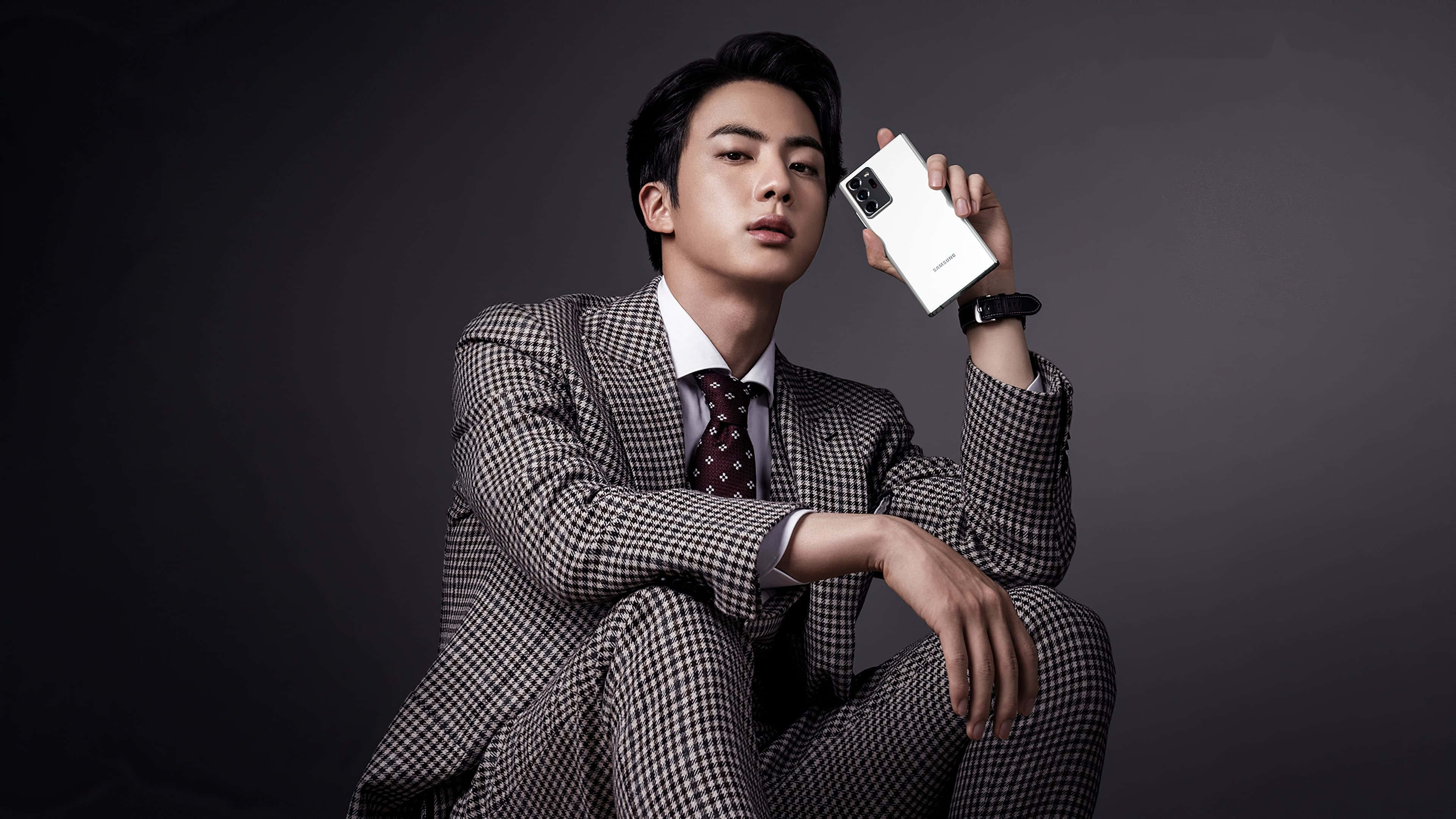 Jin 桌布 壁紙 — 一位穿著格紋西裝的時尚男士，坐在深色攝影棚背景前，手持一部白色智慧型手機。