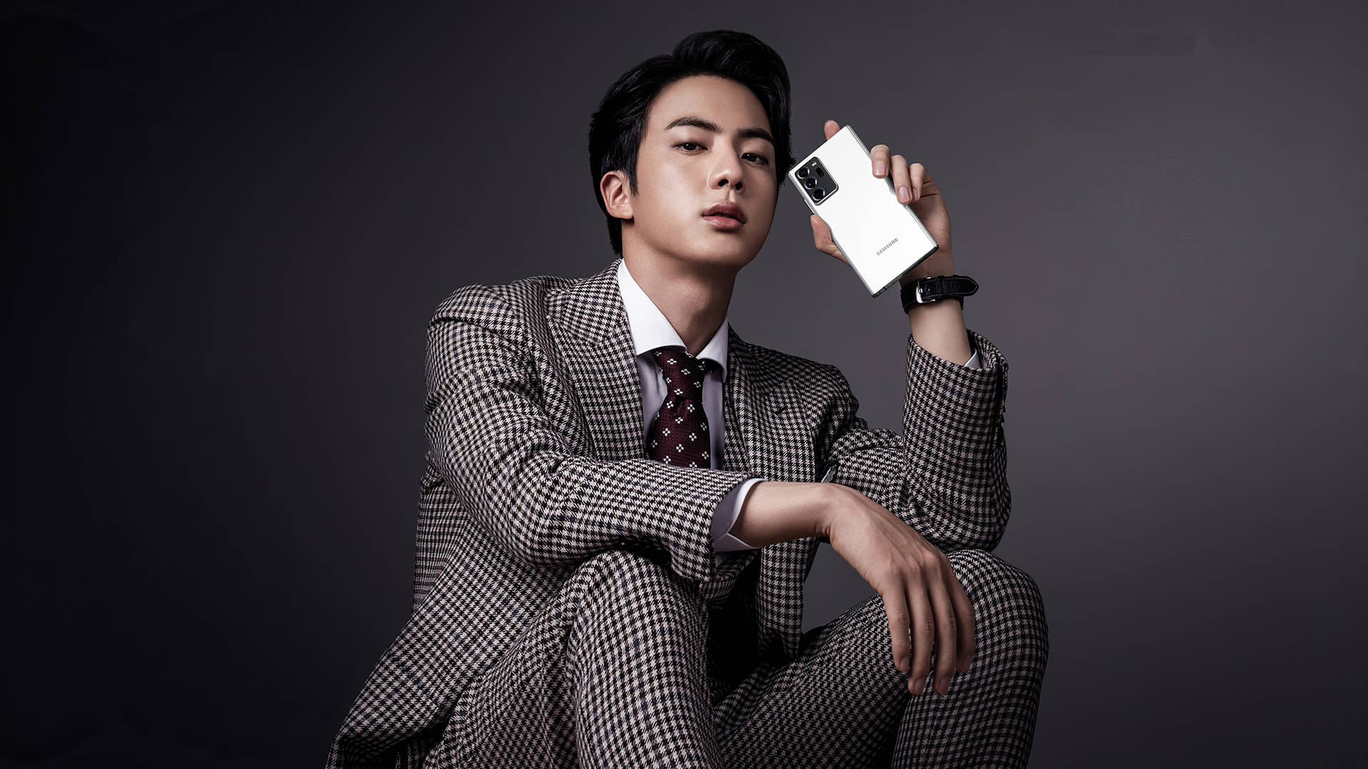 Jin 桌布 壁紙 — 一位穿著格紋西裝的時尚男士在深色攝影棚背景下手持白色智慧型手機。