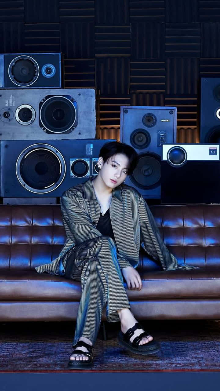 Jungkook 桌布 壁紙 — 一位穿著條紋睡衣的年輕男子，在燈光昏暗的工作室裡，放鬆地坐在一張簇絨皮革沙發上，周圍環繞著復古喇叭。