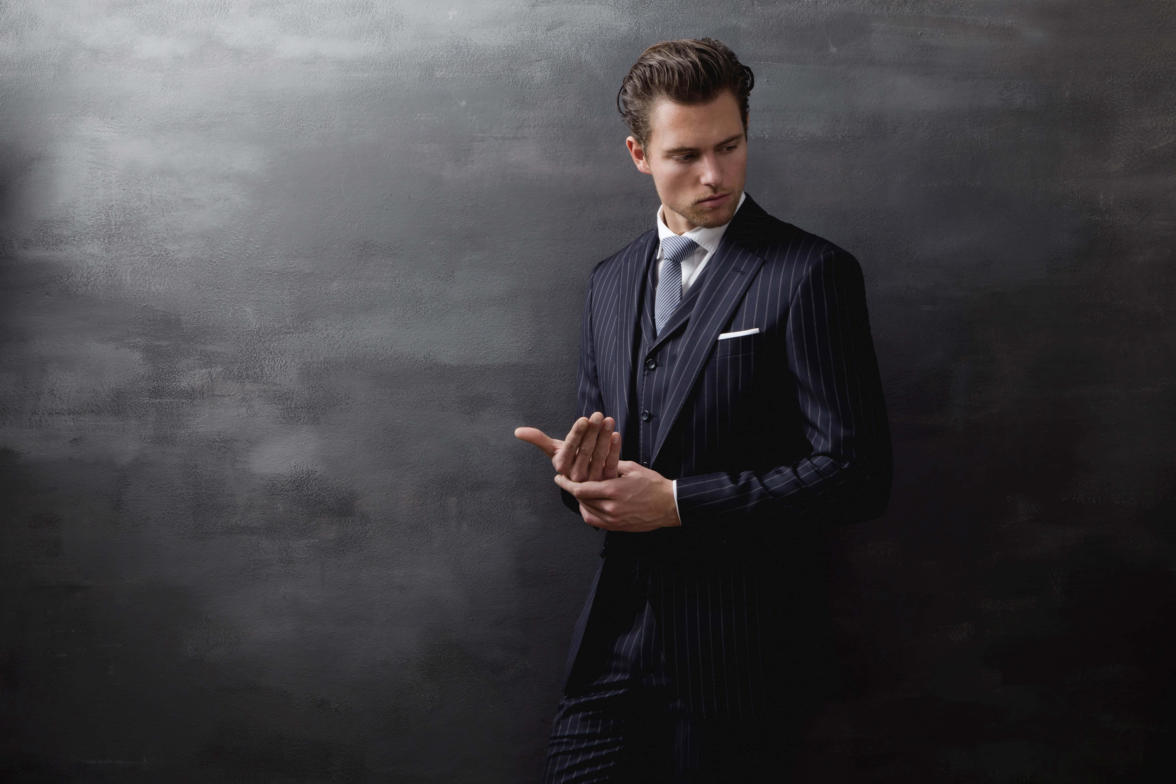 Mens Fashion 桌布 壁紙 — 一位穿著條紋西裝、衣著俐落的男士，在帶有紋理的深灰色背景前用手勢比劃。