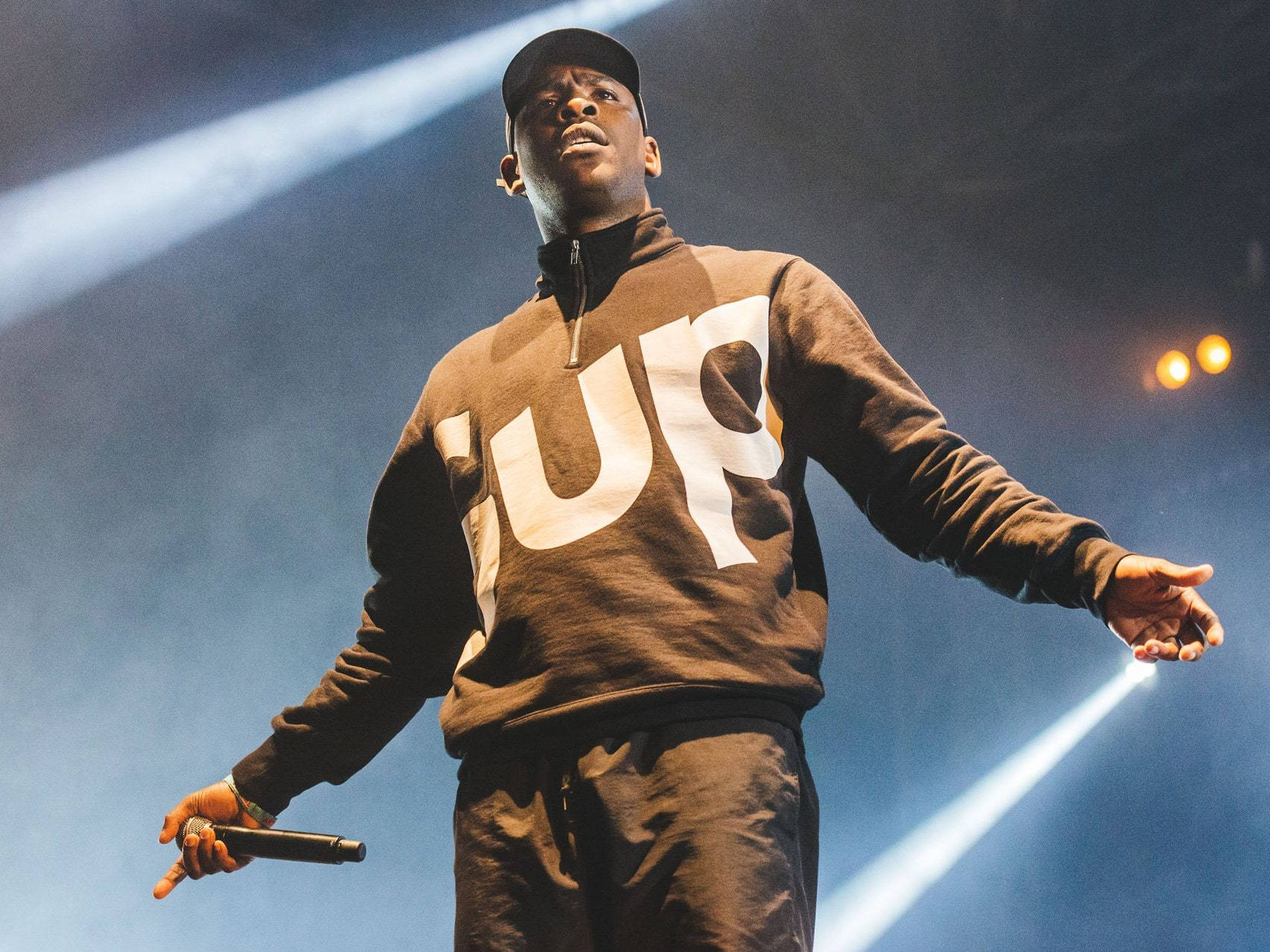 Skepta 桌布 壁紙 — 一位穿著棕色 'SUP' 連帽衫的說唱歌手在明亮的藍色聚光燈下表演，煙霧在他周圍旋轉。