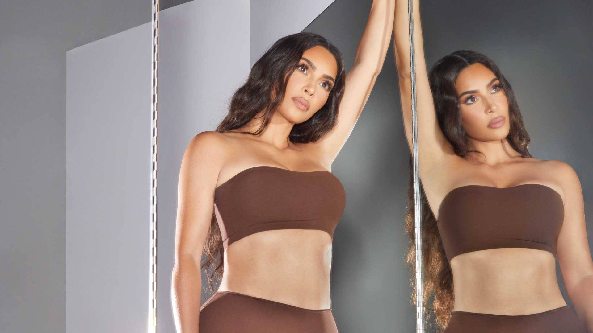 Kim Kardashian 桌布 壁紙 — 一位穿著棕色內衣的女性在極簡灰色牆壁前自信地擺姿勢，她的倒影在鏡中可見。
