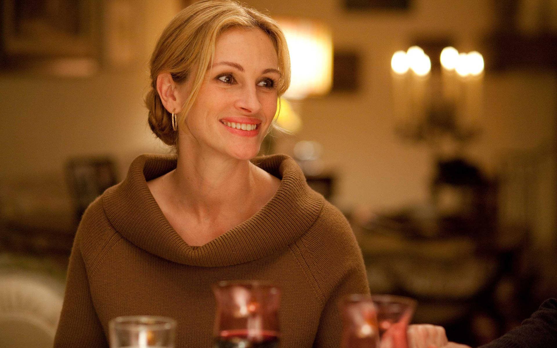 Julia Roberts 桌布 壁紙 — 一位穿著棕色毛衣的微笑女士坐在溫暖燈光的餐桌旁，桌上放著酒杯。