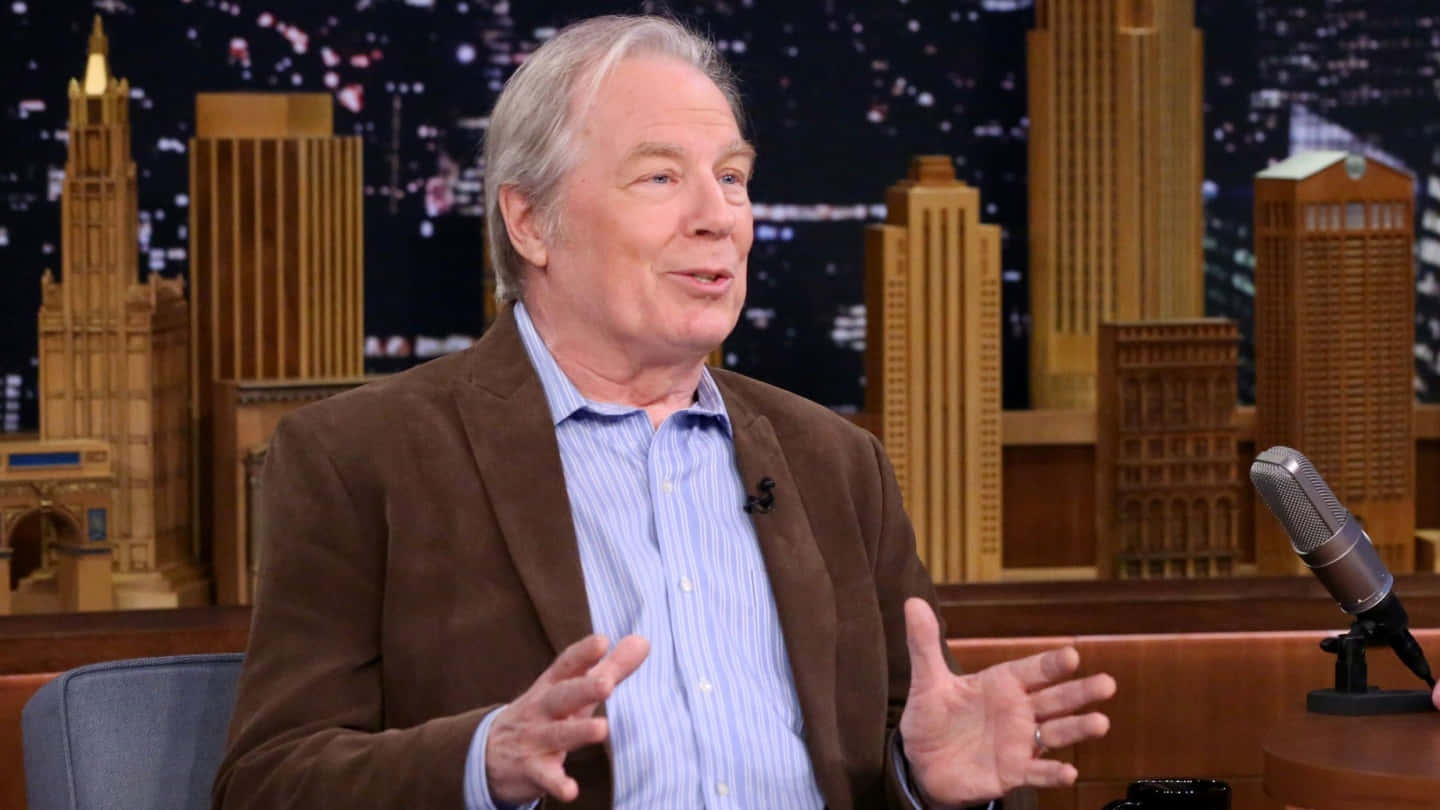 Michael Mckean 桌布 壁紙 — 一位穿著棕色西裝外套的長者，在設有微型城市天際線背景的脫口秀舞台上說話時做手勢。