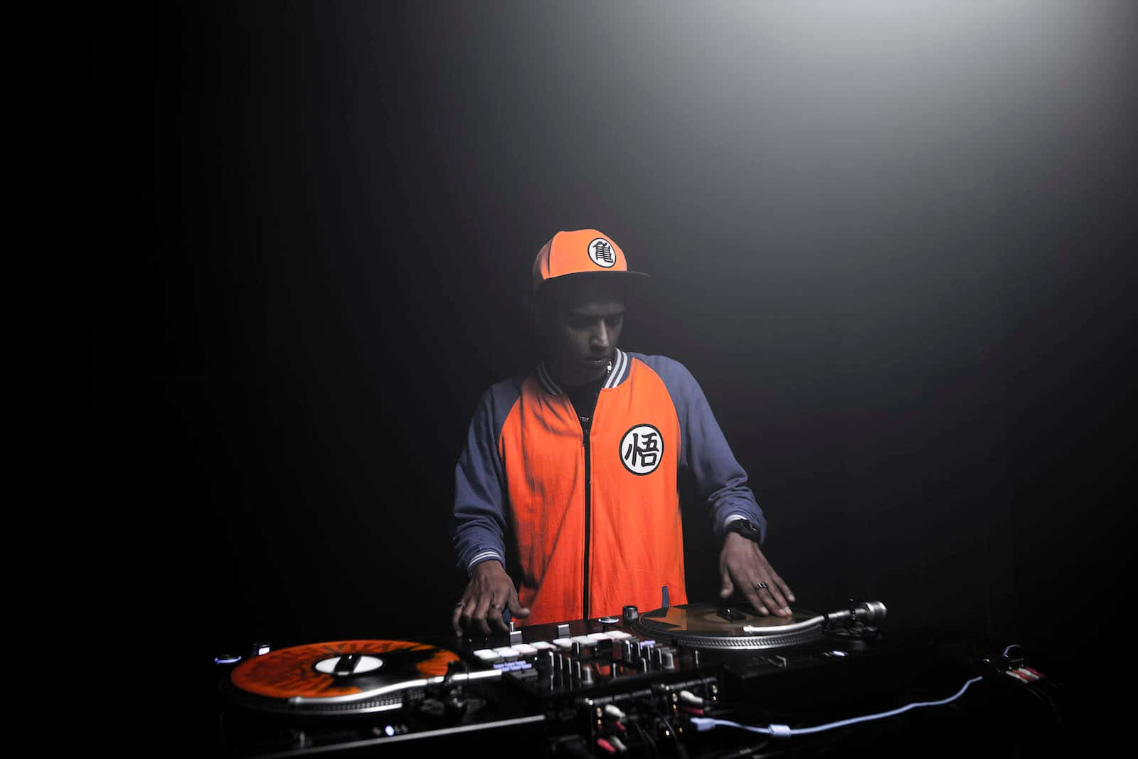 DJ 桌布 壁紙 — 一位穿著橘色悟空外套和帽子的DJ在黑暗背景的聚光燈下混音唱片。