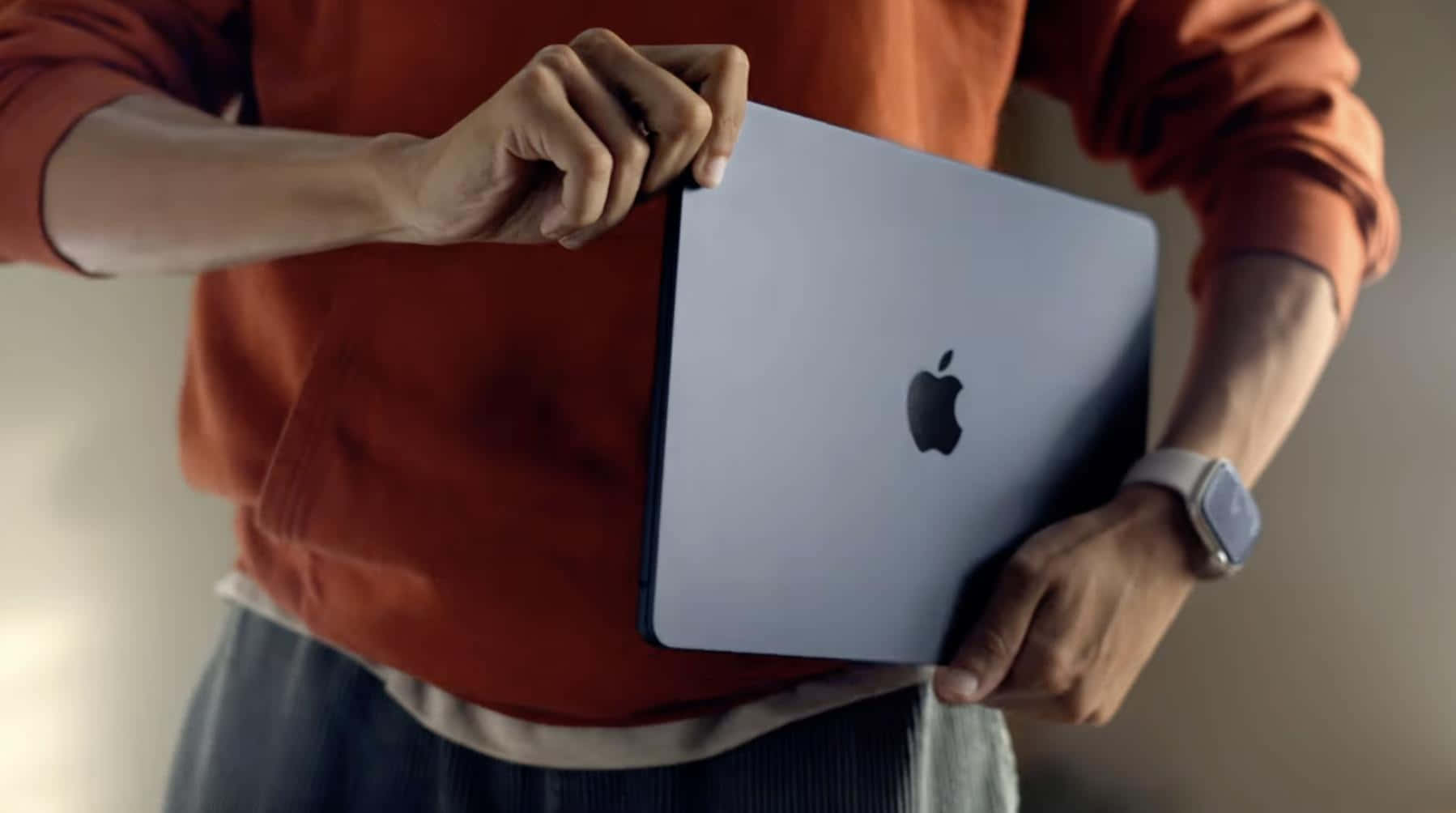 MacBook 動態圖片 桌布 壁紙 — 一位穿著橙色毛衣的人，在柔和的模糊背景前拿著帶有Apple標誌的銀色MacBook。