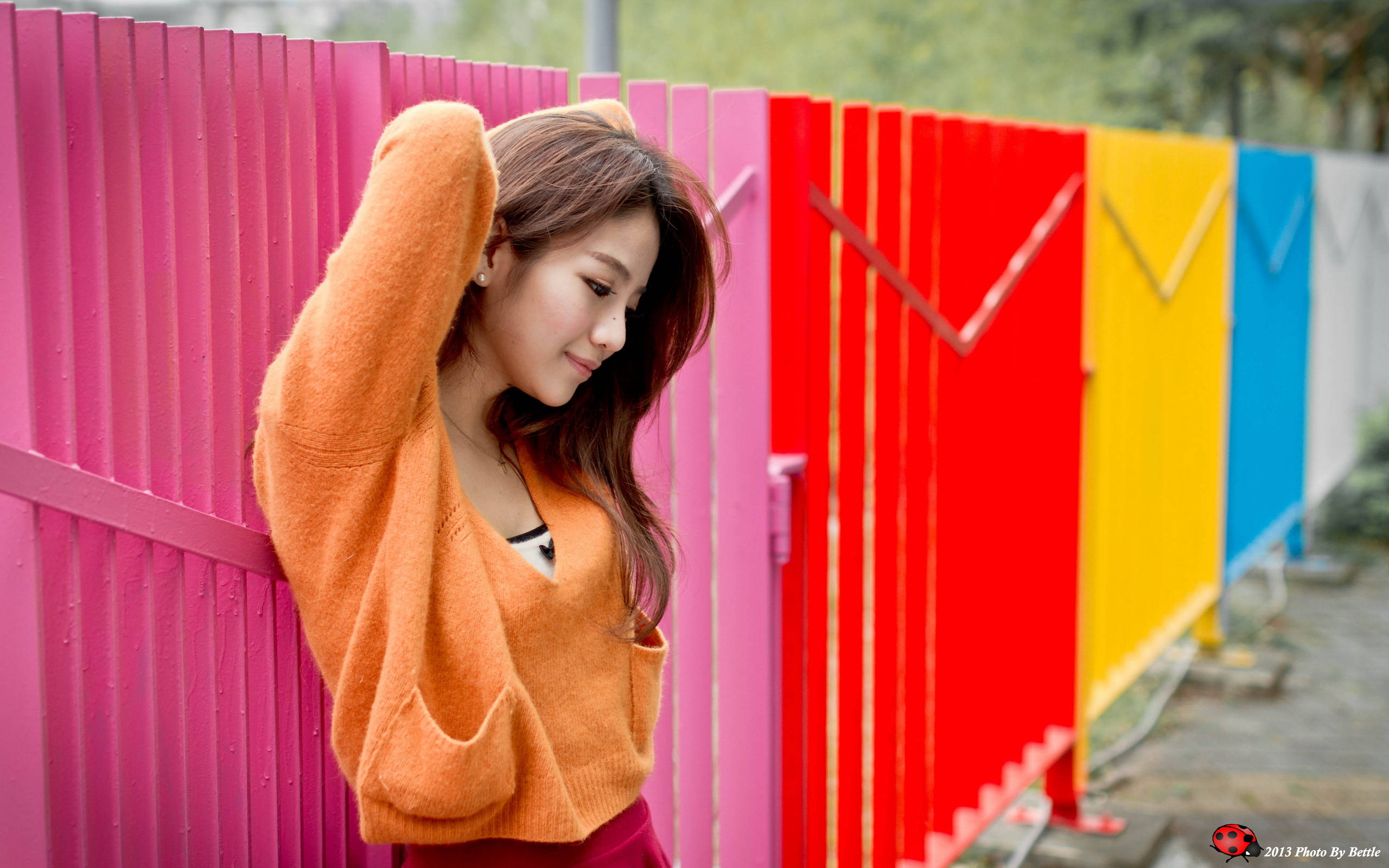 2560x1600 桌布 壁紙 — 一位穿著橙色毛衣的年輕女子，帶著溫柔的微笑，靠著充滿活力的彩虹色圍欄。