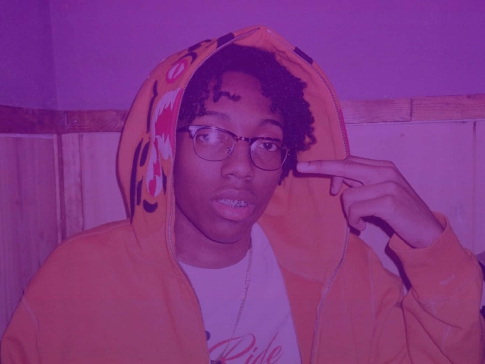 Rappers 桌布 壁紙 — 一位穿著橙色老虎連帽衫和眼鏡的年輕人，在紫色燈光下指著自己的太陽穴。