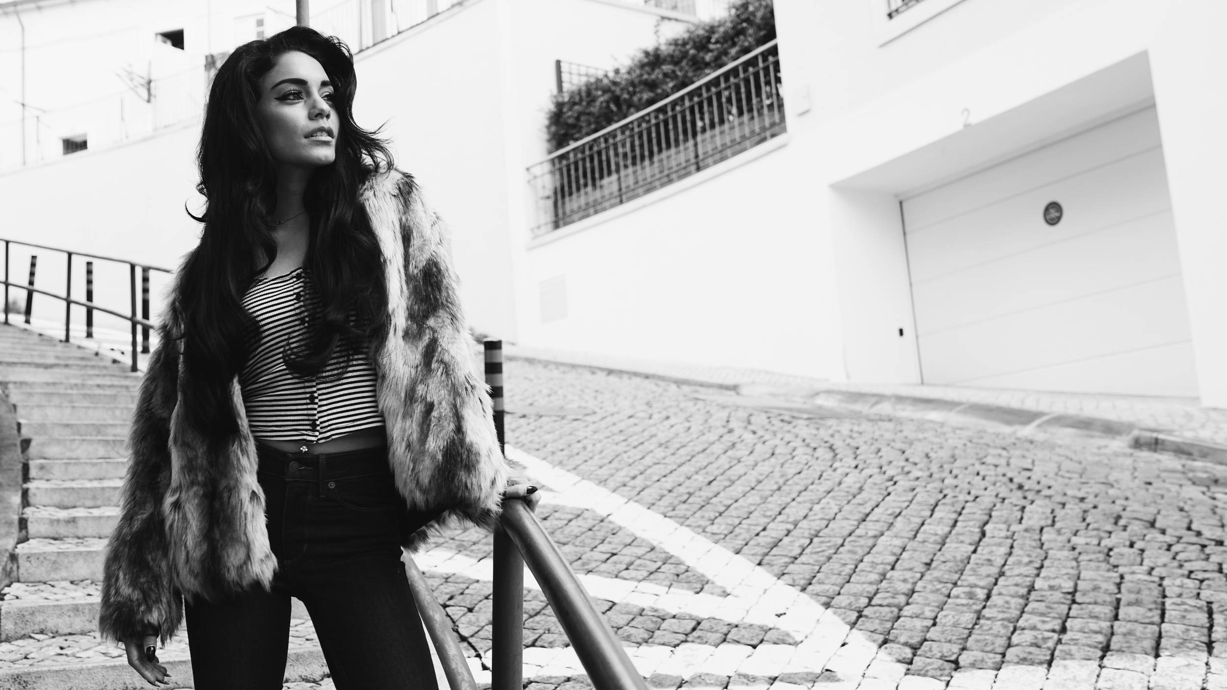 Vanessa Hudgens 桌布 壁紙 — 一位穿著毛皮大衣的女性自信地站在黑白相間的鵝卵石街道上，旁邊是樓梯和一棟白色建築。