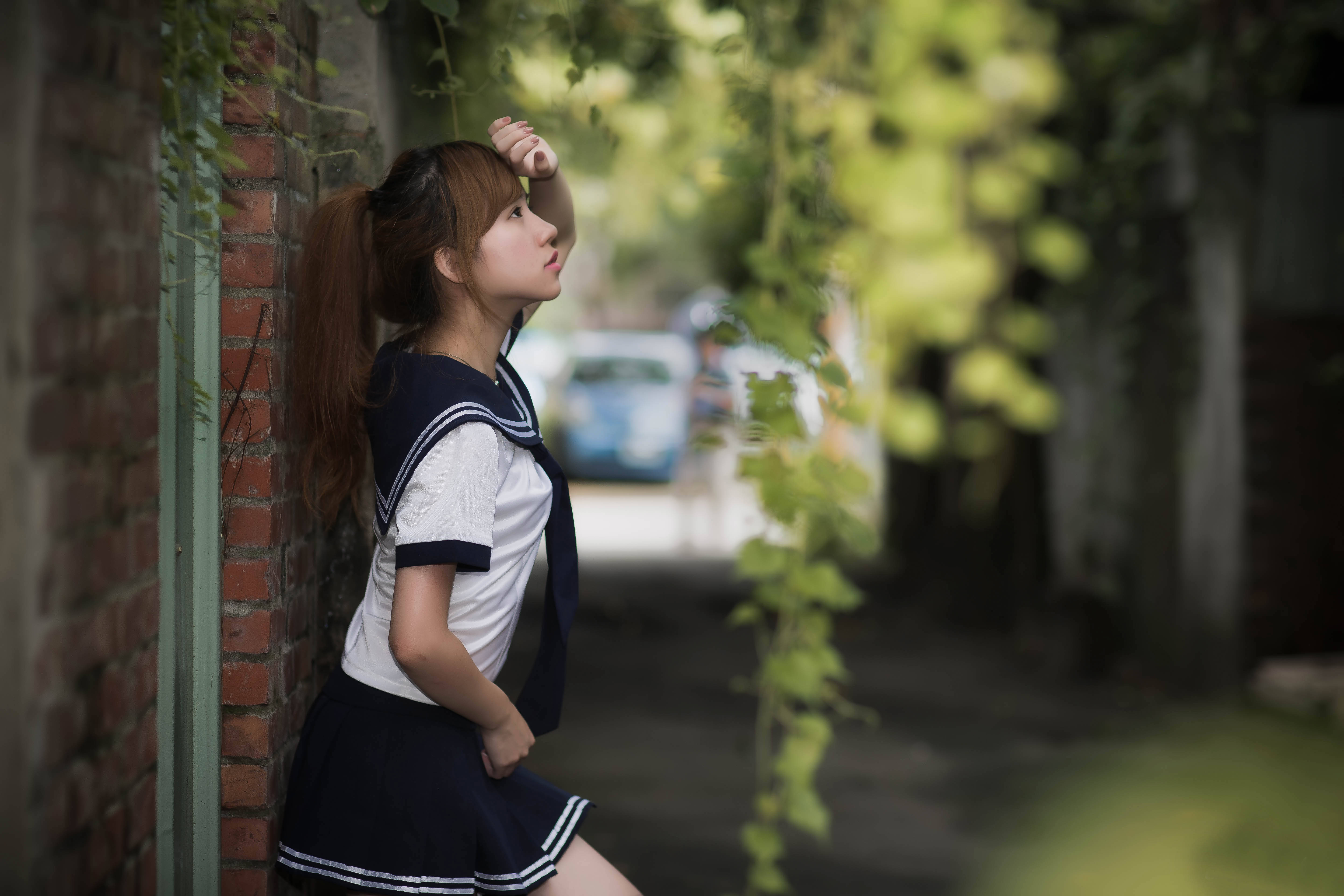 Chinese Girl 桌布 壁紙 — 一位穿著水手服的年輕女子在安靜的小巷裡，靠著磚牆，頭頂是斑駁的綠色樹葉。