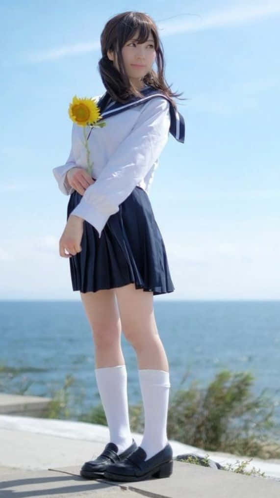 美學 桌布 壁紙 — 一位穿著水手服的年輕女子在晴朗的藍天下，在海邊拿著一朵向日葵。