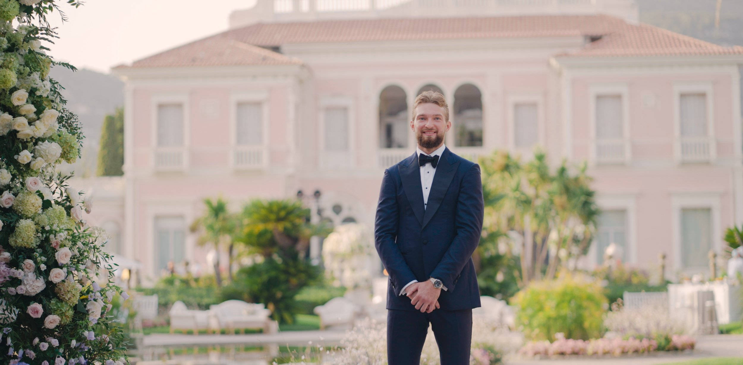domantas sabonis 桌布 壁紙 — 一位穿著海軍藍禮服的新郎面帶微笑，站在一座宏偉的粉紅色別墅前，周圍環繞著茂盛的花園和花卉佈置。