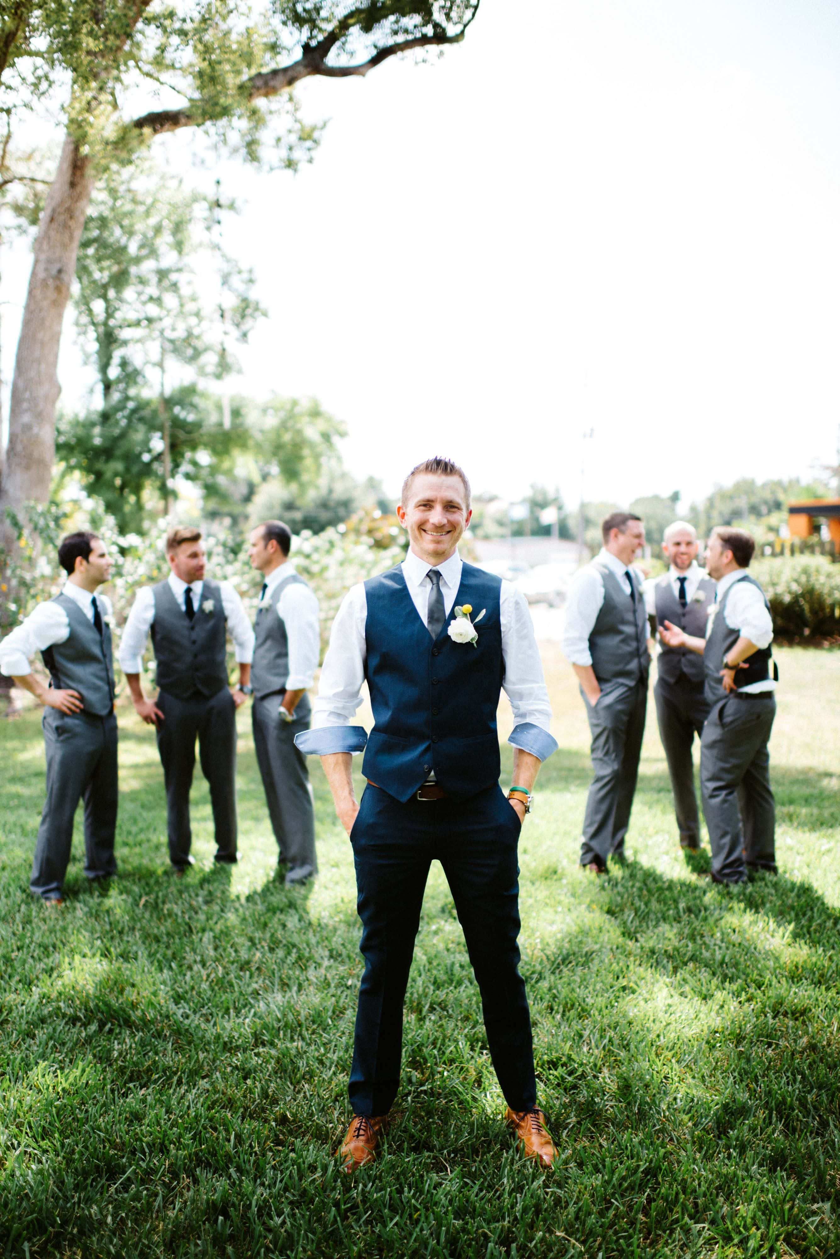 Groomsmen 桌布 壁紙 — 一位穿著海軍藍背心和棕色鞋子的新郎對著鏡頭微笑，他的伴郎們穿著配套的西裝站在他身後，在陽光明媚的草地上。
