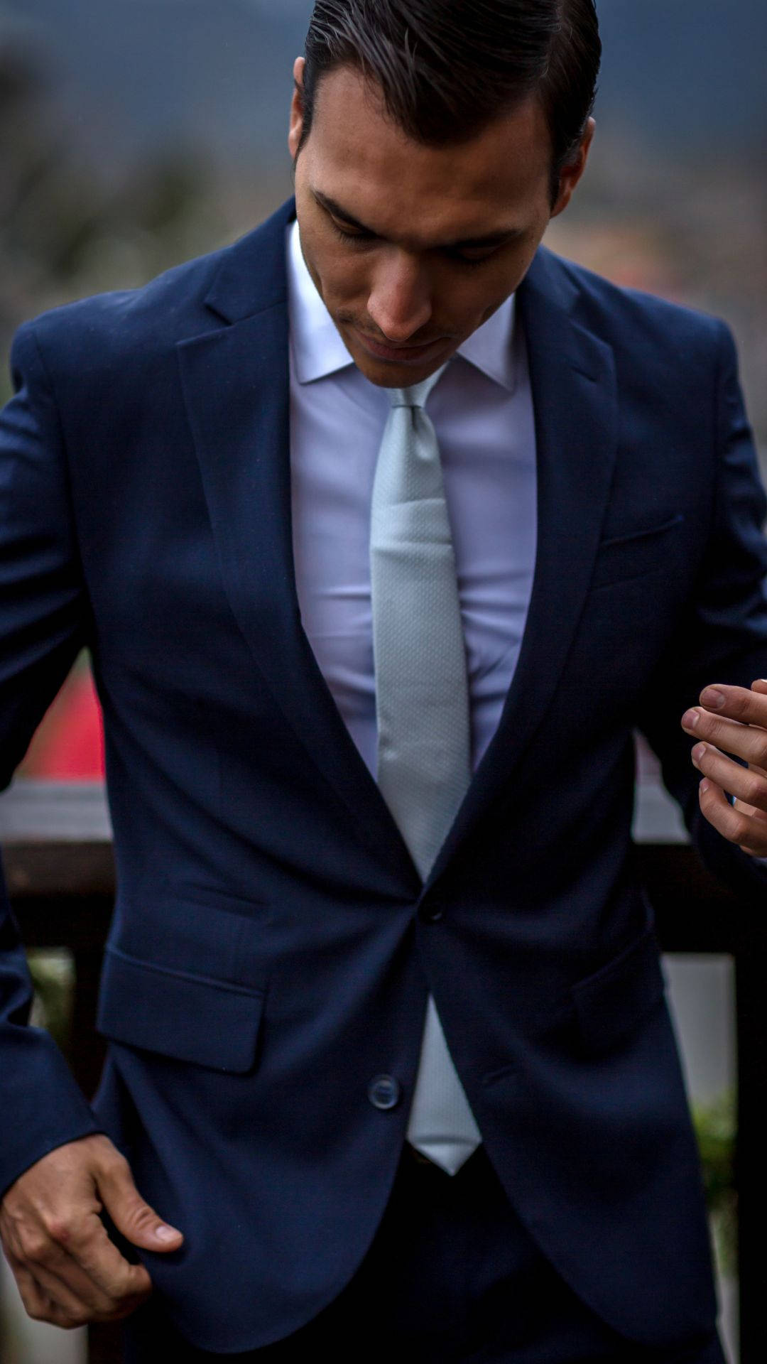 suit 桌布 壁紙 — 一位穿著海軍藍西裝和淺色領帶、衣著講究的男子，在模糊的戶外背景下低頭，表情嚴肅。
