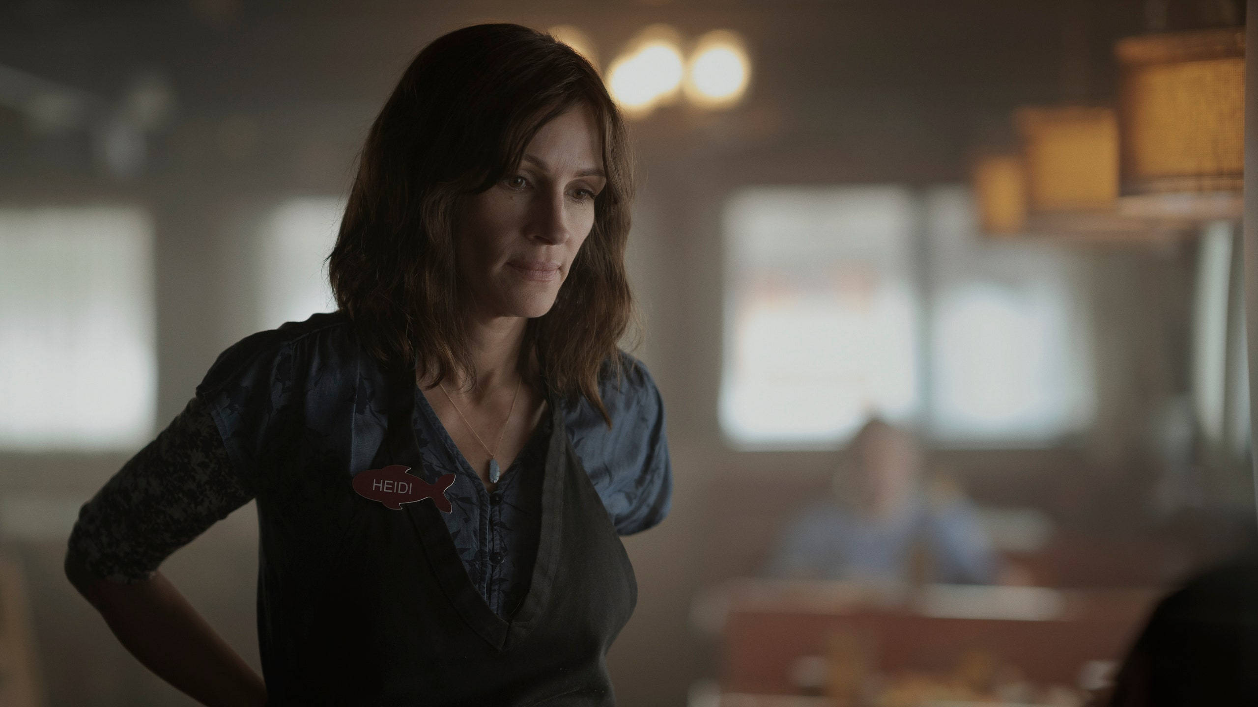 Julia Roberts 桌布 壁紙 — 一位穿著深色圍裙並帶有名牌的女性，站在一家燈光溫暖、背景帶有柔和散景效果的餐廳裡。
