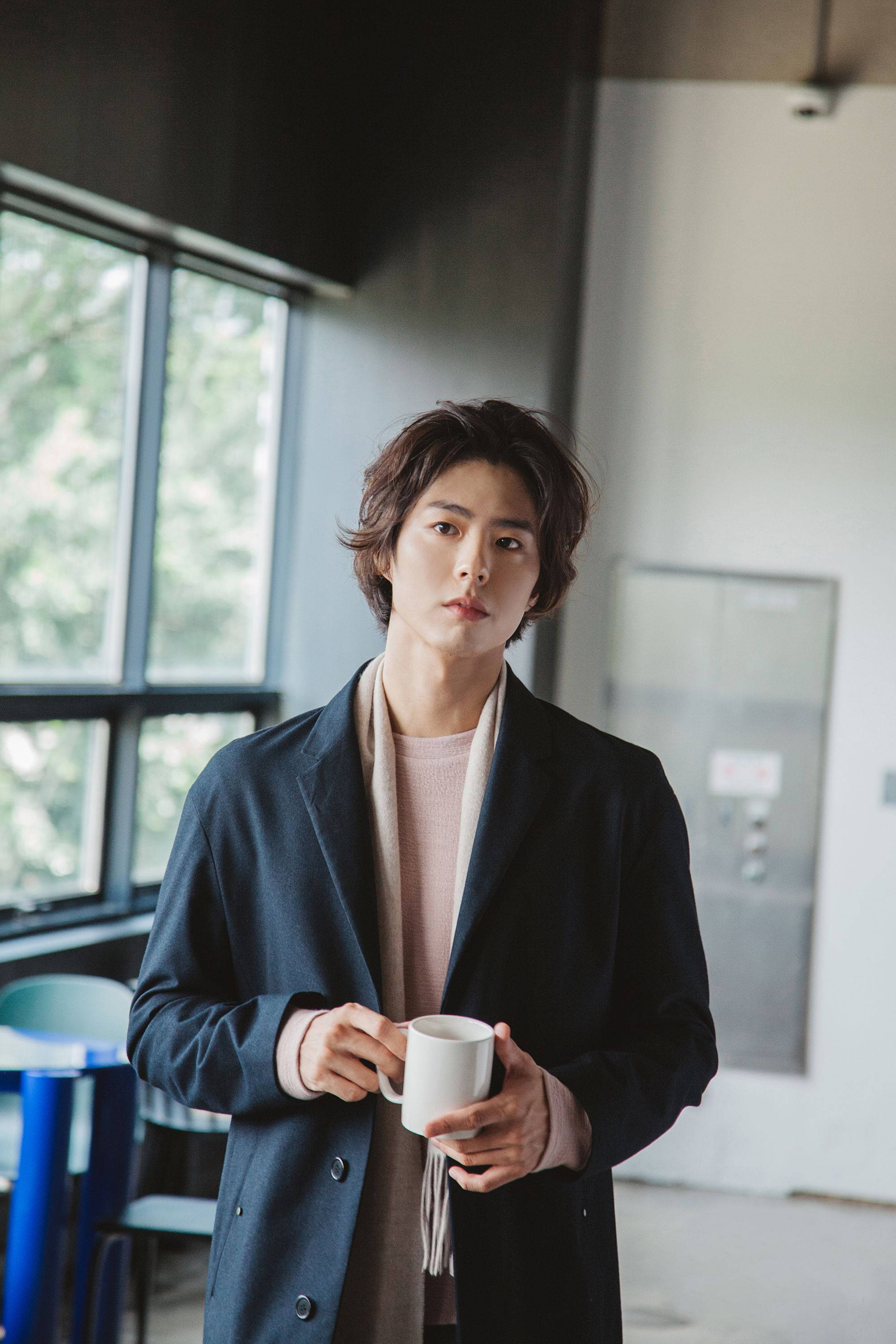 Reply 1988 桌布 壁紙 — 一位穿著深色外套的年輕男子，在現代室內空間靠近窗戶的地方，手持一個白色馬克杯。