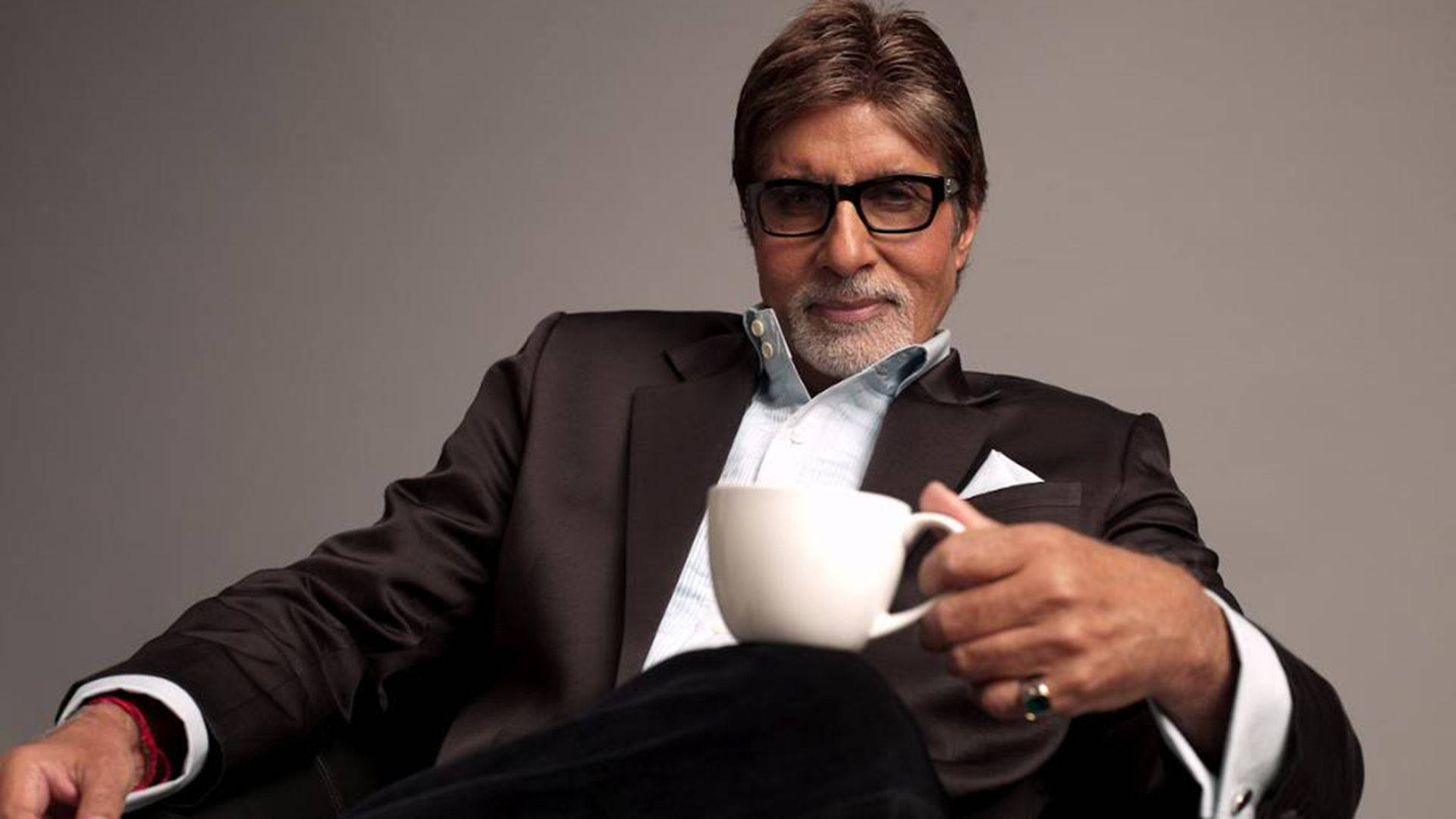 Amitabh Bachchan 桌布 壁紙 — 一位穿著深色西裝、戴眼鏡的尊貴男士，坐在中性灰色背景前，手持白色咖啡杯。