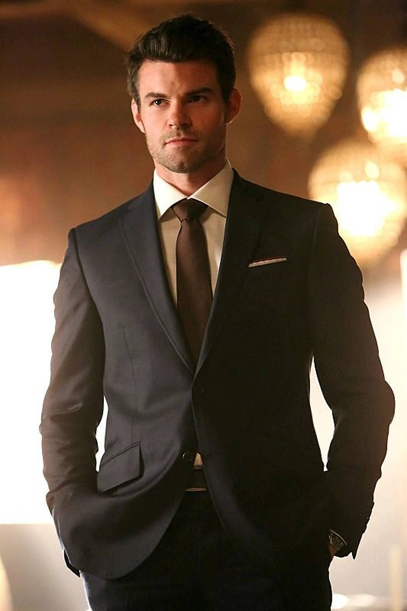 elijah mikaelson 桌布 壁紙 — 一位穿著深色西裝、打扮俐落的男士，雙手插袋，自信地站在溫暖燈光下、模糊的閃爍水晶吊燈背景前。