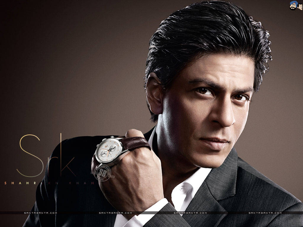 Shahrukh 桌布 壁紙 — 一位穿著深色西裝、打扮俐落的男士，在柔和的工作室燈光下，於低調的棕色背景前手持一只奢華手錶。