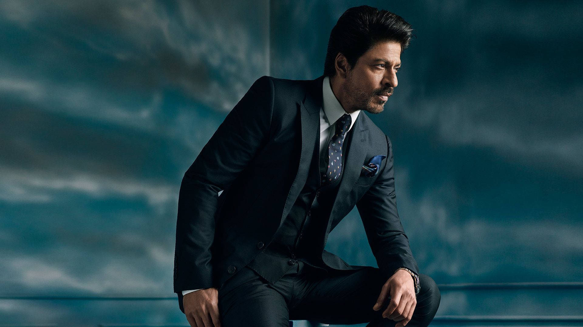 Shahrukh 桌布 壁紙 — 一位穿著深色西裝、打扮俐落的男士，自信地坐在陰鬱的青色天空背景下。