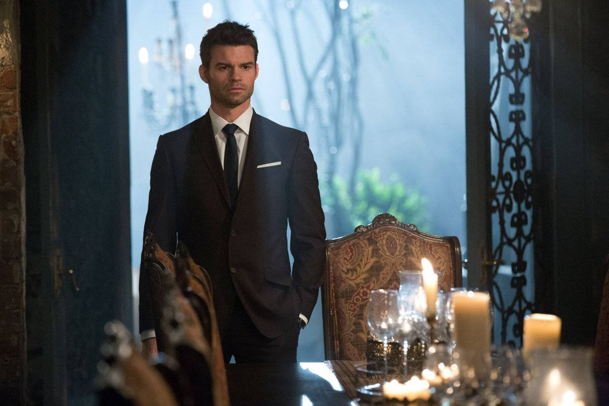 elijah mikaelson 桌布 壁紙 — 一位穿著深色西裝、衣著講究的男子站在光線昏暗的飯廳裡，周圍有華麗的椅子和閃爍的蠟燭。