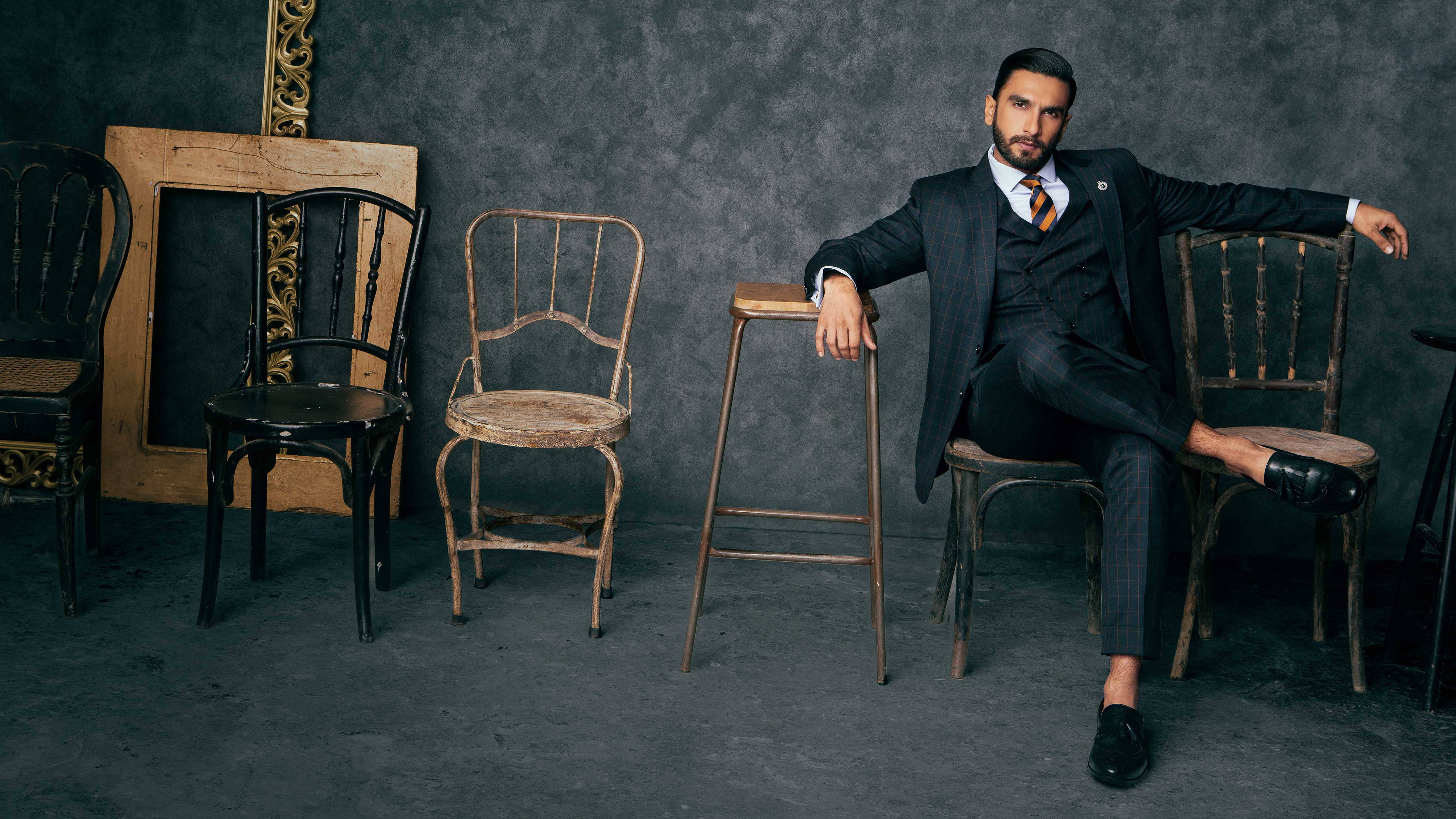 Ranveer Singh 桌布 壁紙 — 一位穿著深色西裝、衣著講究的男子，在質感灰色牆壁前，在一堆復古椅子中的一張鄉村風格椅子上自信地閒坐。