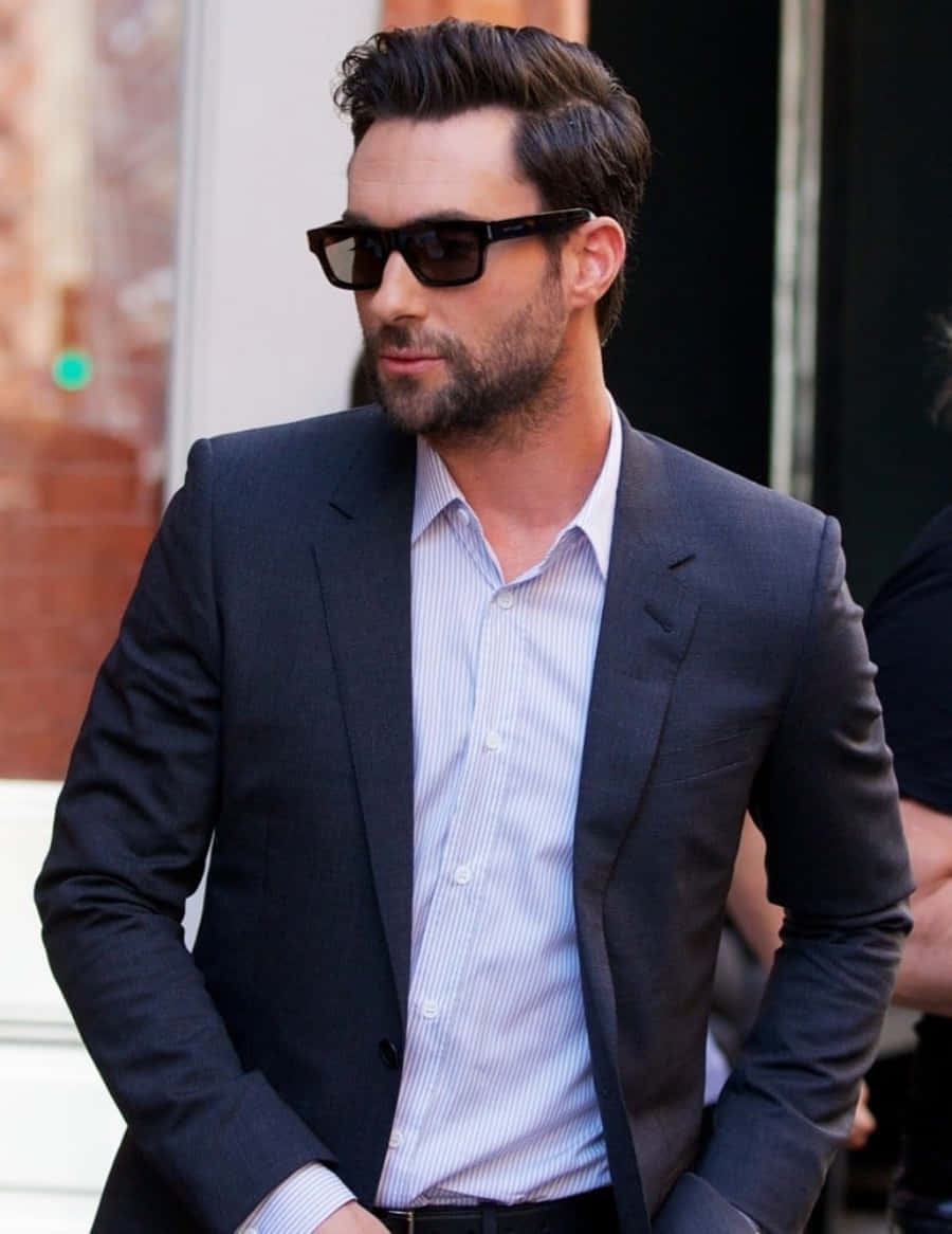 Adam Levine 桌布 壁紙 — 一位穿著深色西裝和太陽眼鏡的時尚男士，自信地走在城市街道上，背景是磚砌建築。