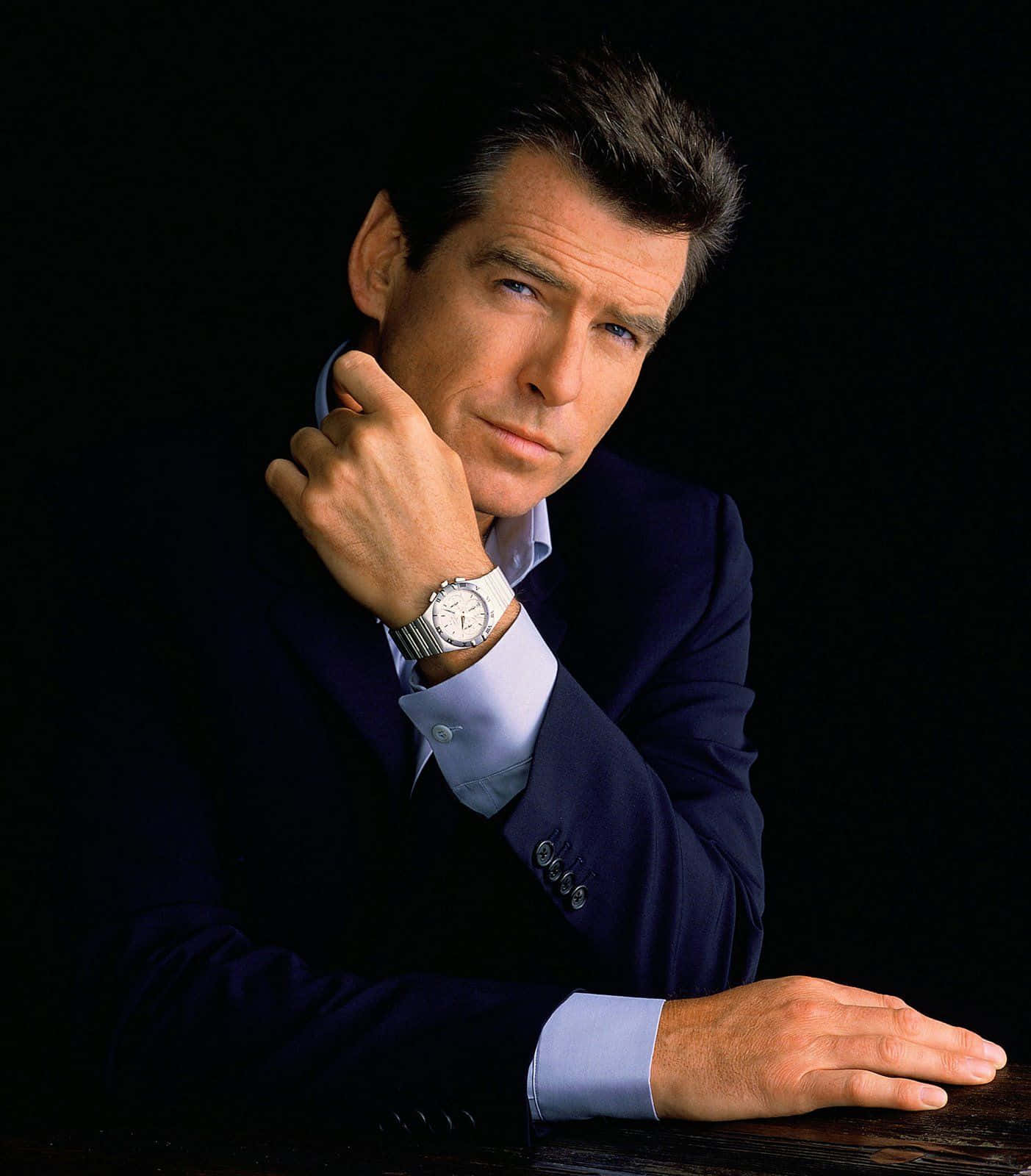 pierce brosnan 桌布 壁紙 — 一位穿著深色西裝和淺色襯衫、打扮俐落的男士，在黑色背景前自信地擺姿勢，手托下巴，手腕上可見一隻銀色手錶。