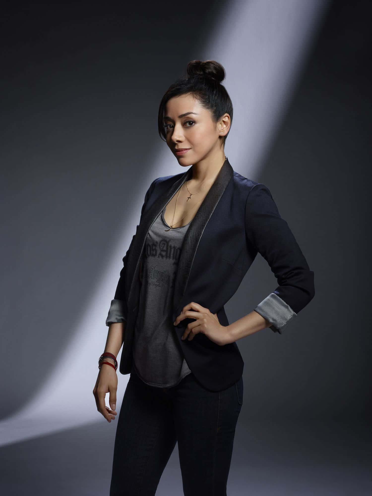 Aimee Garcia 桌布 壁紙 — 一位穿著深色西裝外套和牛仔褲的優雅女性，在灰色工作室背景的戲劇性聚光燈下自信地站立。