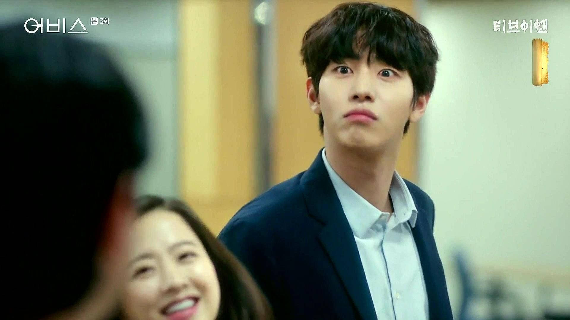 Ahn Hyo Seop 桌布 壁紙 — 一位穿著深色西裝外套的年輕男子睜大眼睛驚訝地凝視，而一位微笑的女子坐在他旁邊，處於溫暖照明的室內環境中。