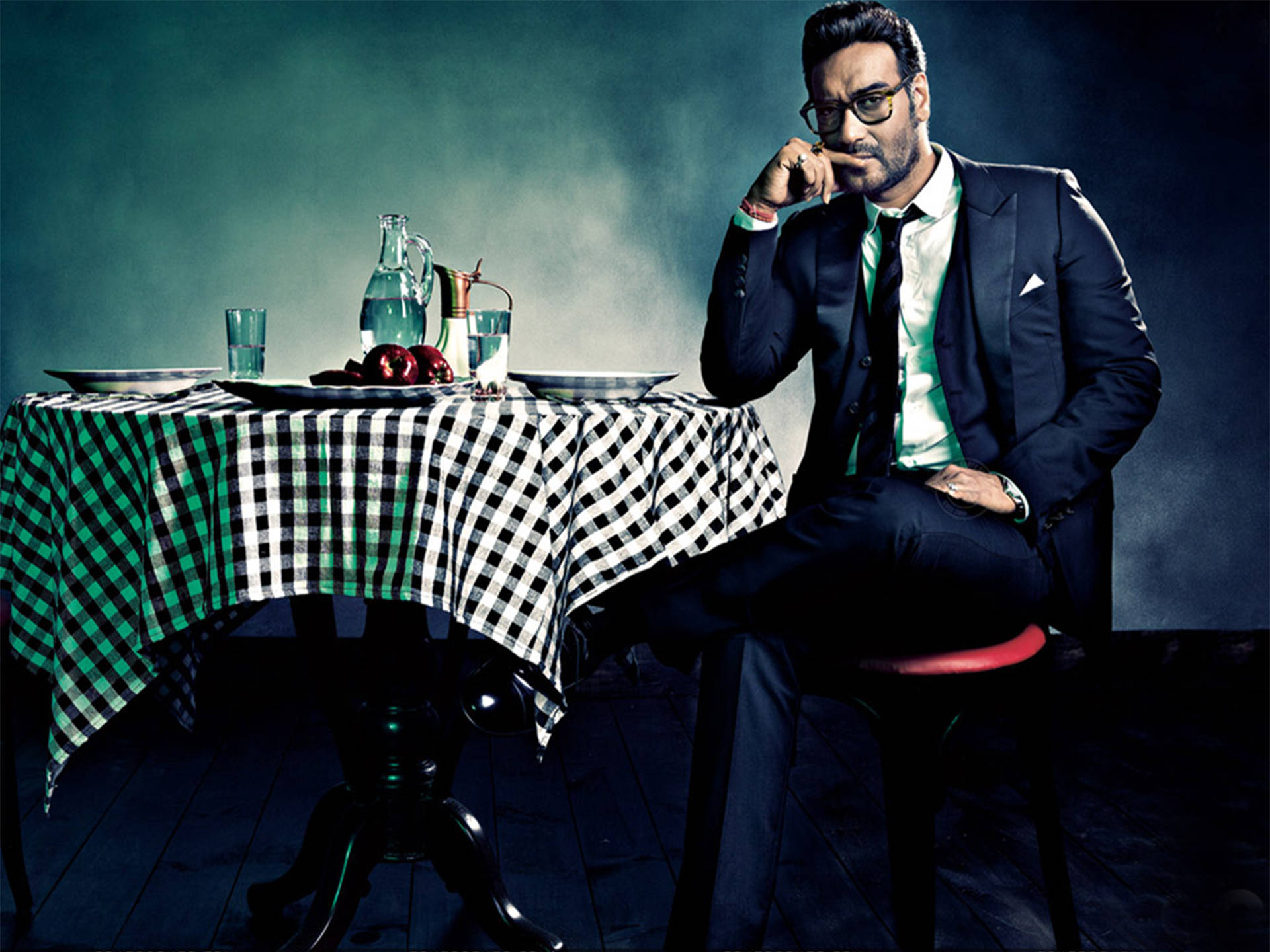 ajay devgn 桌布 壁紙 — 一位穿著深色西裝的時尚男士，在陰暗、燈光昏暗的攝影棚環境中，坐在鋪著格子桌布的桌子旁沉思。