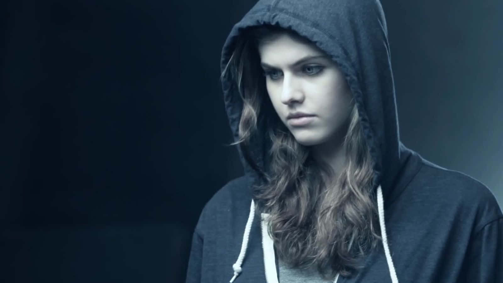 Alexandra Daddario 桌布 壁紙 — 一位穿著深色連帽衫的年輕女子，在柔和的藍色背景下，帶著嚴肅的表情專注地凝視。
