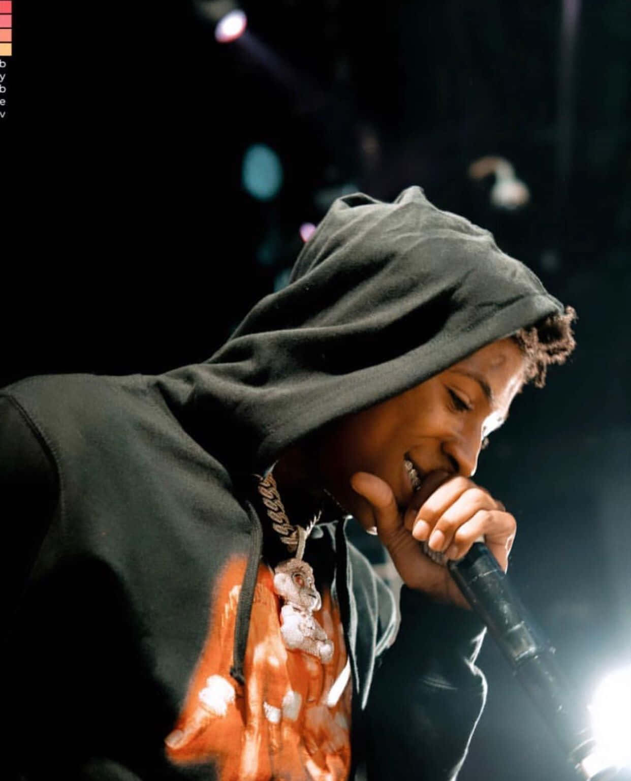 Youngboy 桌布 壁紙 — 一位穿著深色連帽衫的年輕表演者，在明亮的舞台燈光下對著麥克風唱歌時微笑。