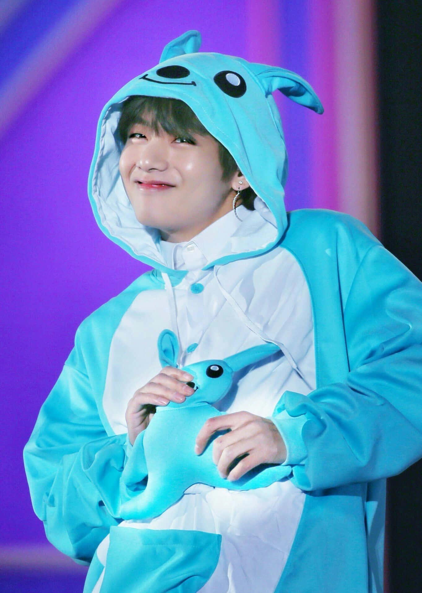 BTS 桌布 壁紙 — 一位穿著淺藍色兔子連身衣的表演者，在充滿活力的紫色舞台背景下，溫暖地微笑著，同時抱著一個相配的毛絨玩具。