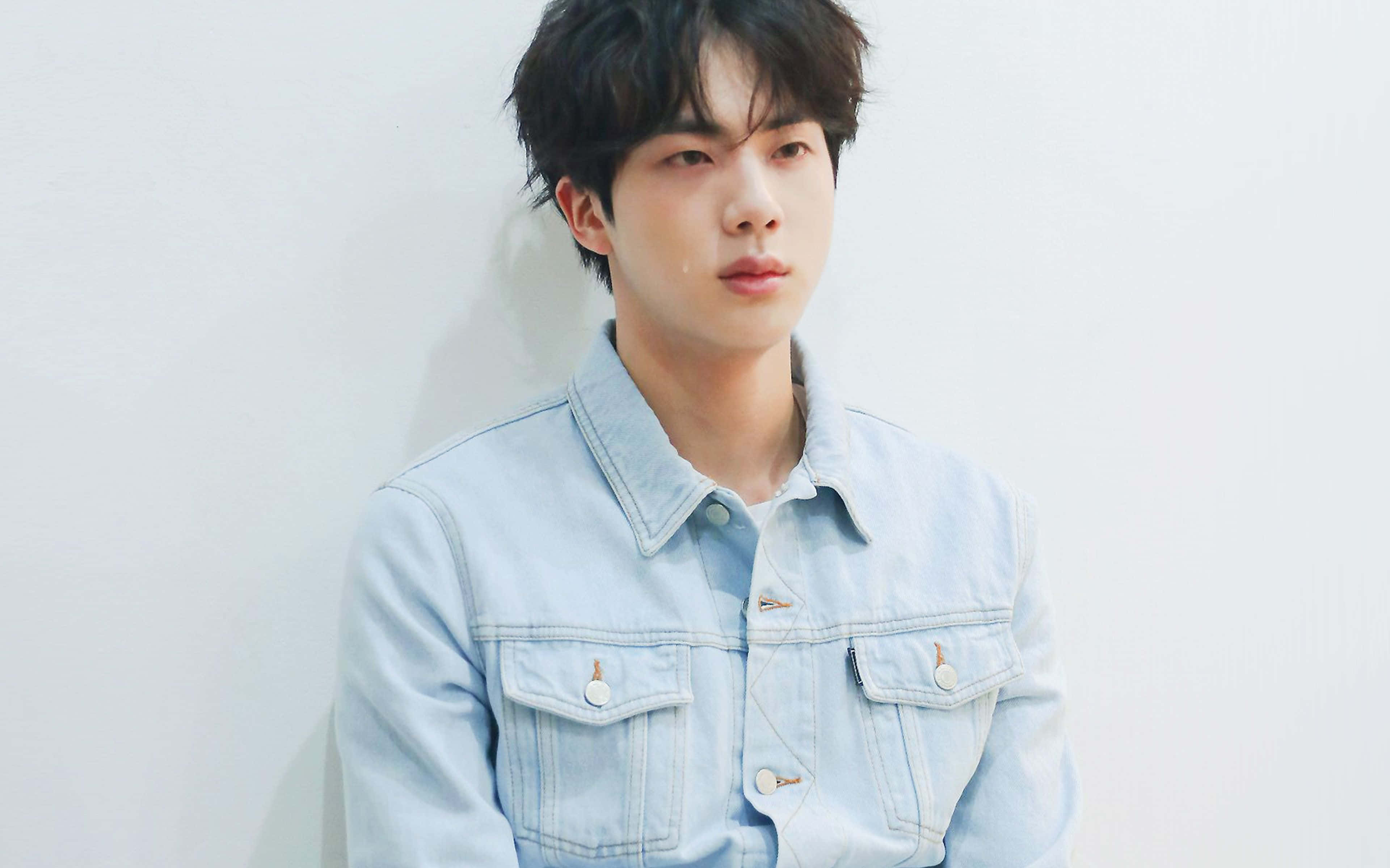 Bts Mac 桌布 壁紙 — 一位穿著淺藍色牛仔外套的年輕男子，在柔和自然光線下靠著白色牆壁。