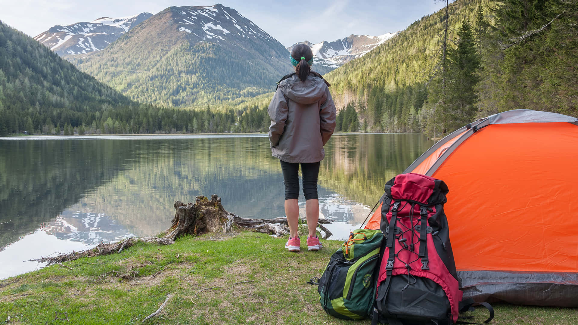 Camping 桌布 壁紙 — 一位穿著灰色外套的遠足者站在一個鮮橙色帳篷旁，俯瞰著一個被雪山和茂密森林環繞的寧靜湖泊。