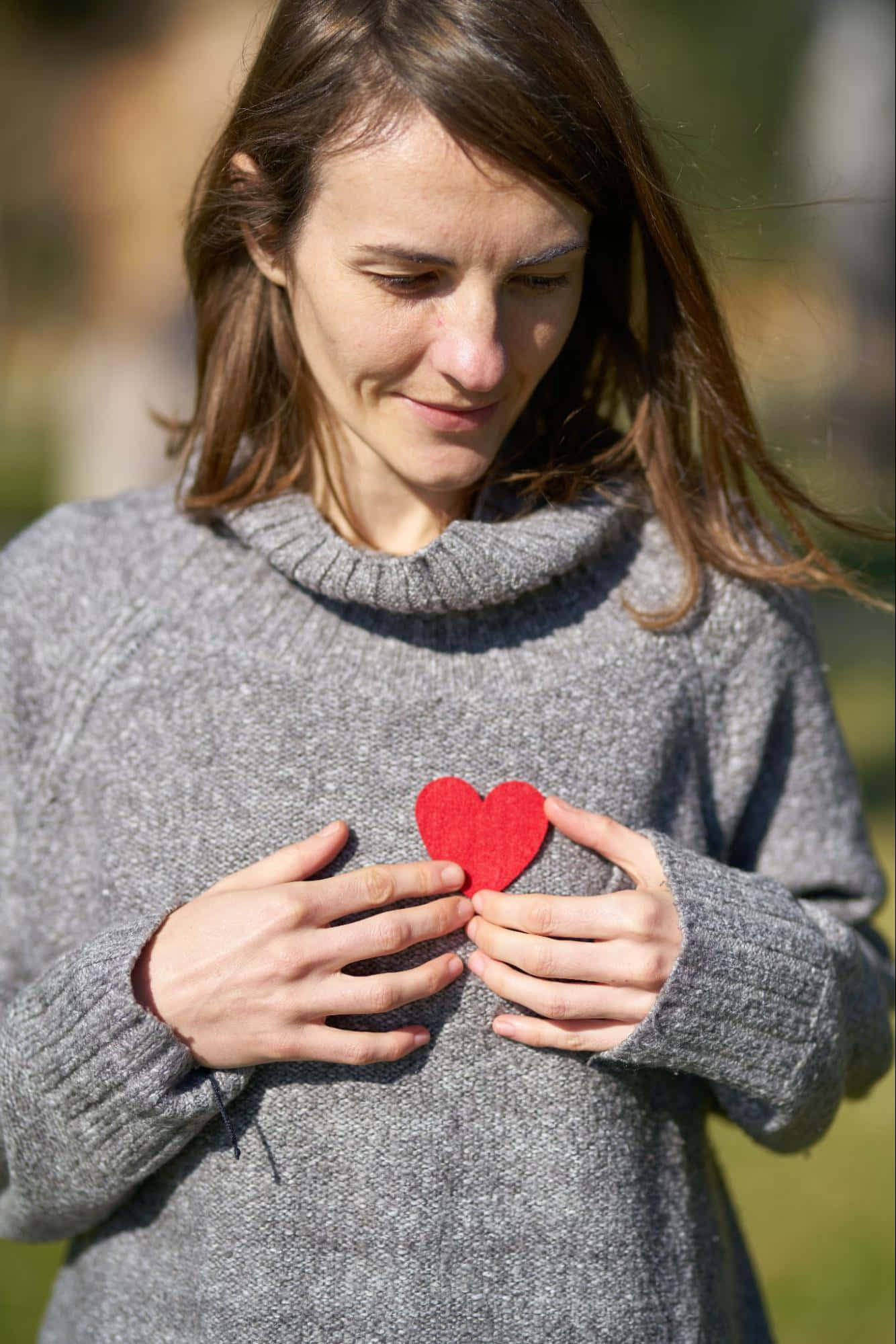 Woman 桌布 壁紙 — 一位穿著灰色毛衣的女性在陽光明媚的戶外環境中，輕輕地將一個紅色氈心放在胸前。