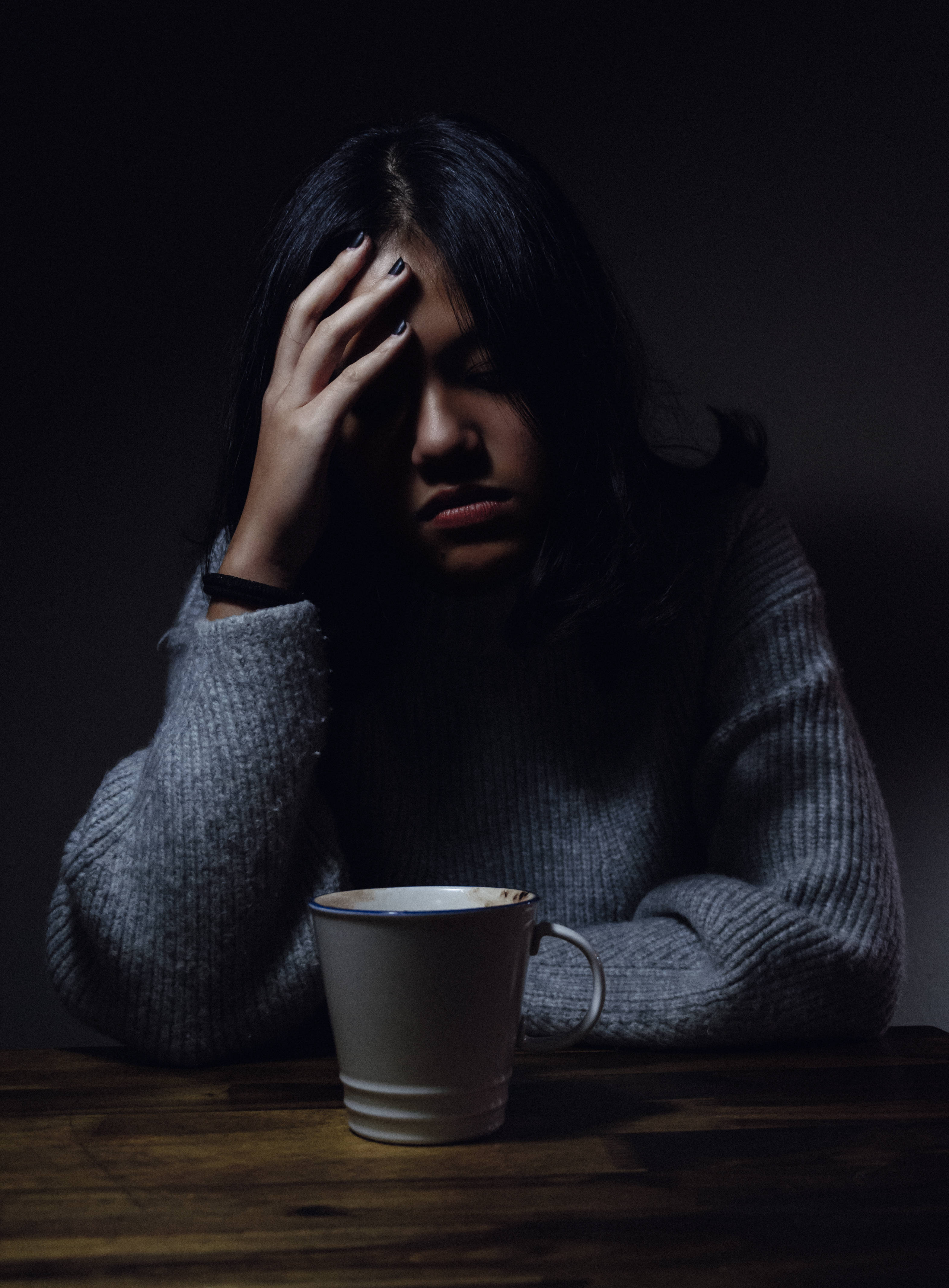 Pain 桌布 壁紙 — 一位穿著灰色毛衣的女性坐在木桌旁，在昏暗的燈光下，用手托著頭，旁邊放著一個白色馬克杯。